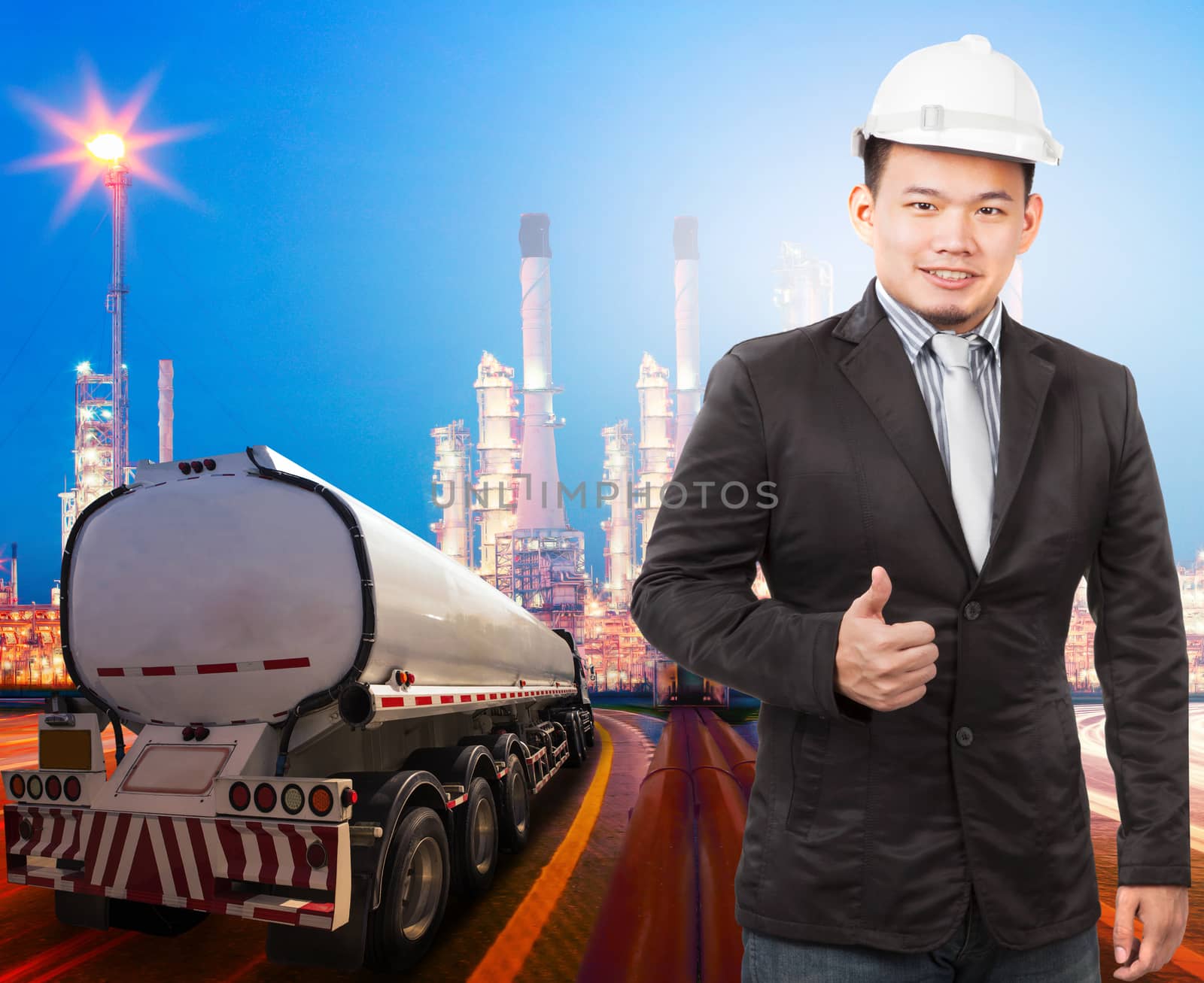 young engineering man with safety helmet standing against beautiful lighting of oil refinery plant in heavy petrochemical industry and container truck transportation of petroleum