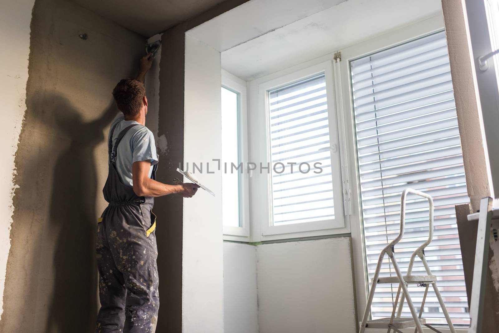 Construction worker wearing worker overall with wall plastering tools renovating apartment house. Plasterer renovating indoor walls and ceilings with float and plaster. Construction finishing works.