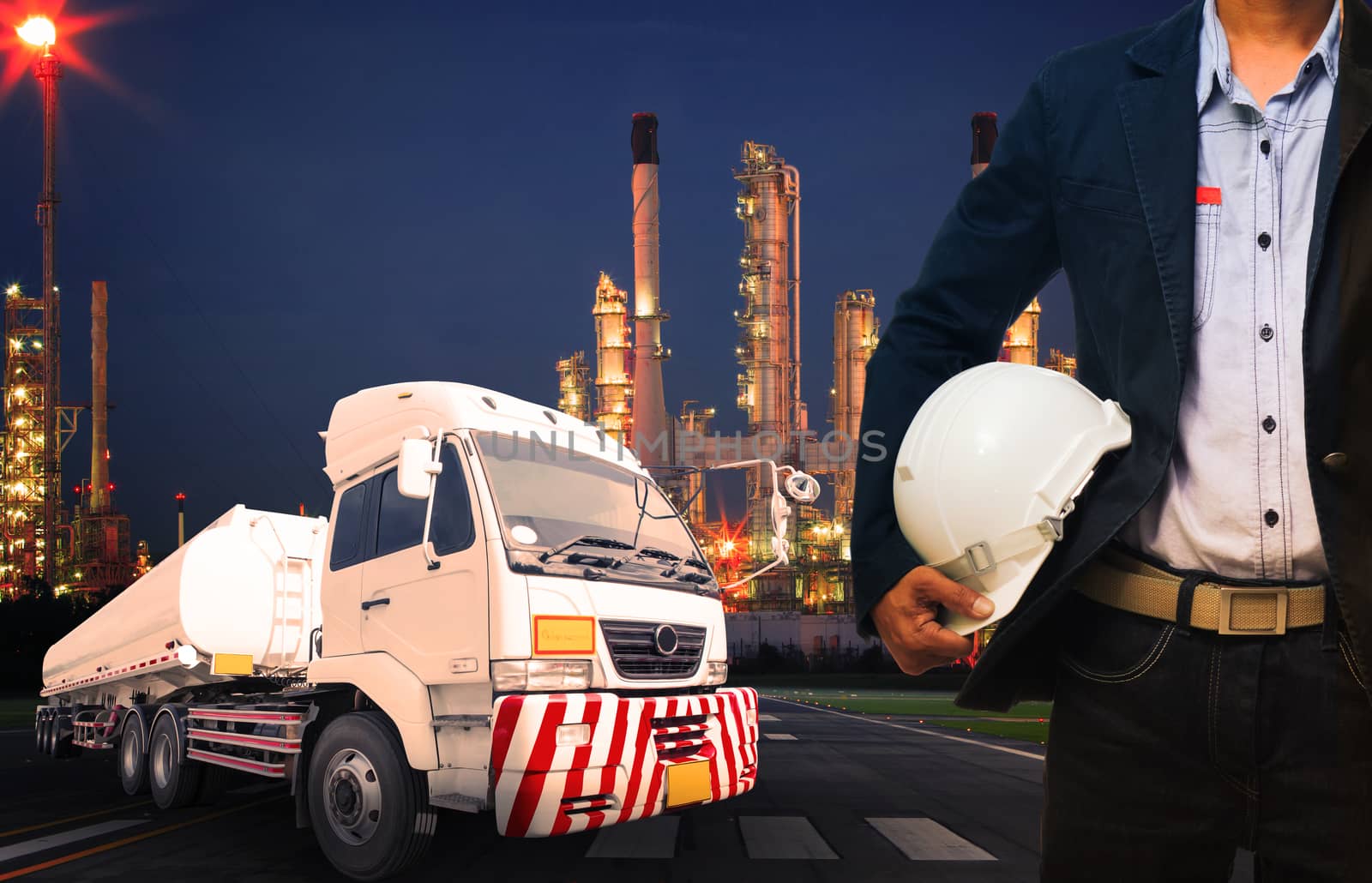 engineer holding hard hat standing against beautiful lighting of oil refinery in heavy petrochemical industry and big oil container truck use for petroleum industrial and land transport business