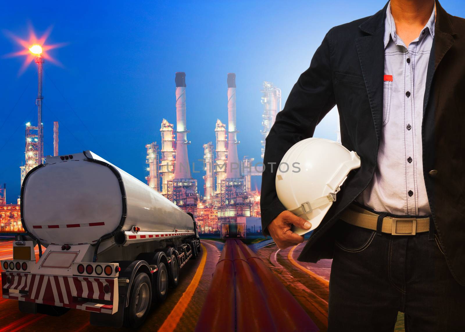 engineering man with safety helmet standing against beautiful lighting of oil refinery plant in heavy petrochemical industry and container truck transportation of petroleum