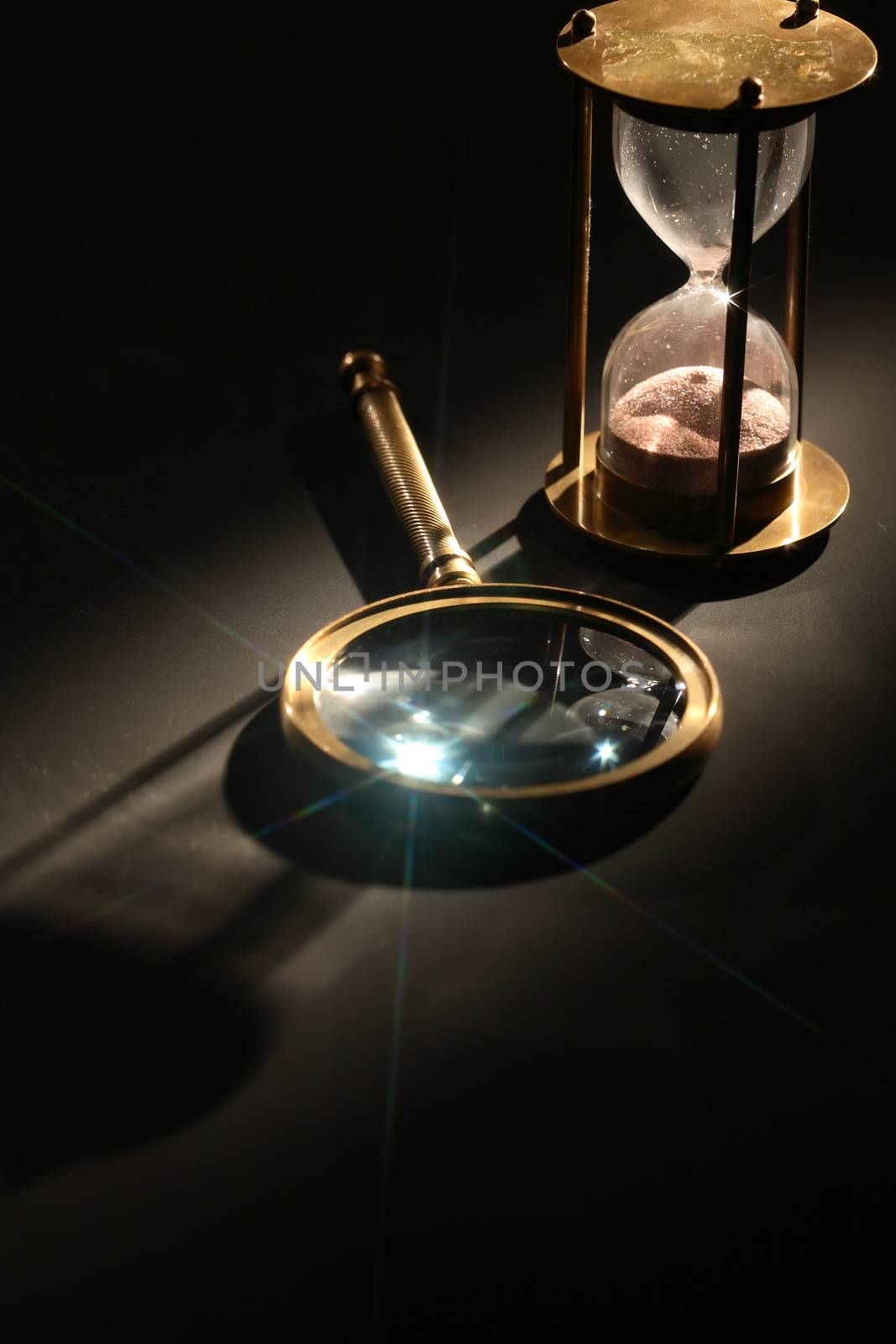 Vintage hourglass near magnifying glass on dark background with long shadow