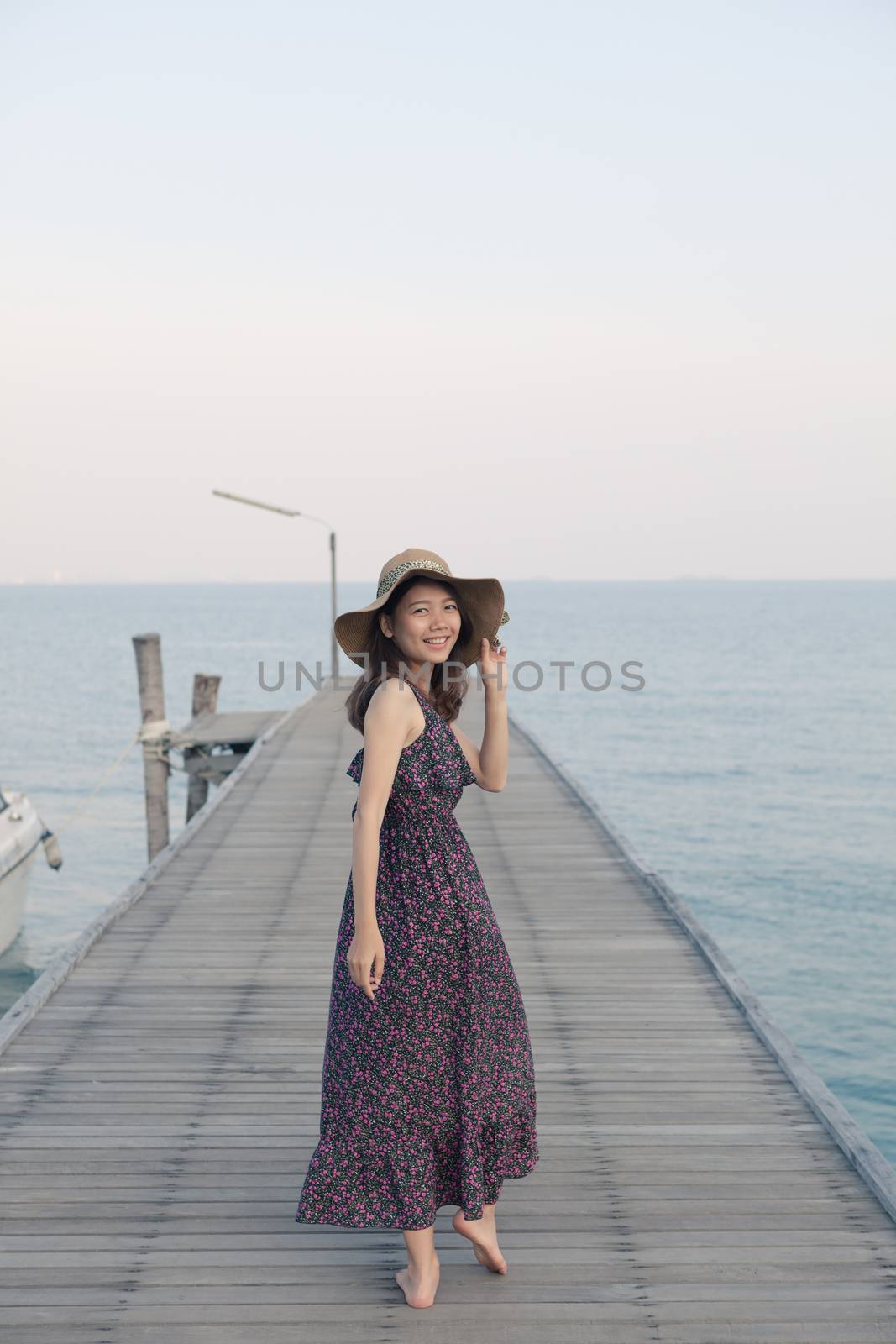 portrait of beautiful young woman wearing wide straw hat and long dress standing with happiness emotion on piers at sea beach 