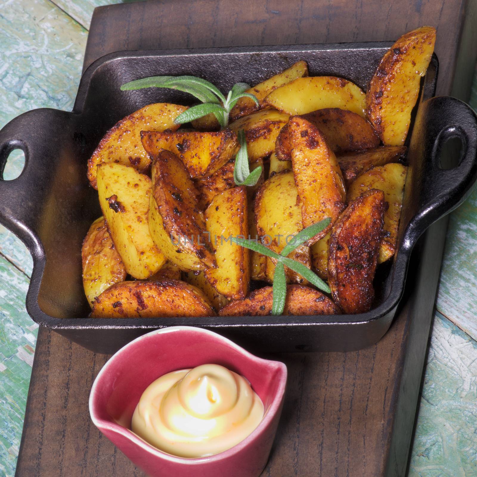 Delicious Roasted Potato Wedges with Rosemary in Black Cast-Iron Pan and Cheese Sauce on Wooden Plate closeup