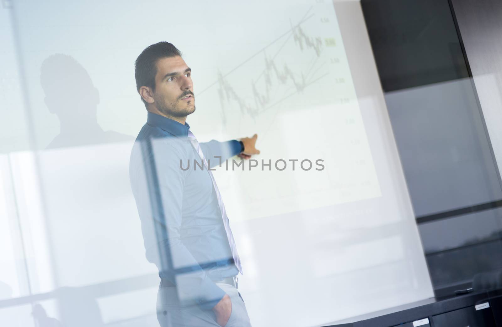 Business man making a presentation in front of whiteboard. Business executive delivering a presentation to his colleagues during meeting or in-house business training. View through glass.