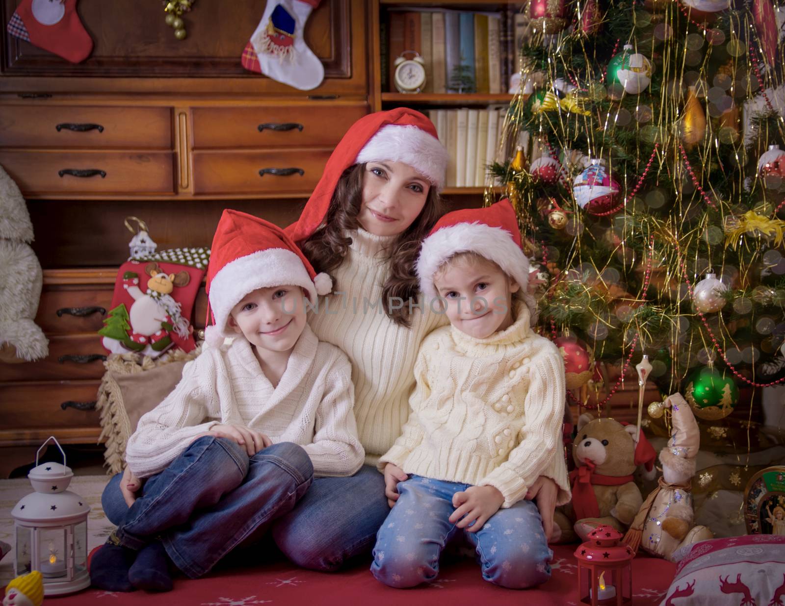 Two children sitting with mother under Christmas tree in hats