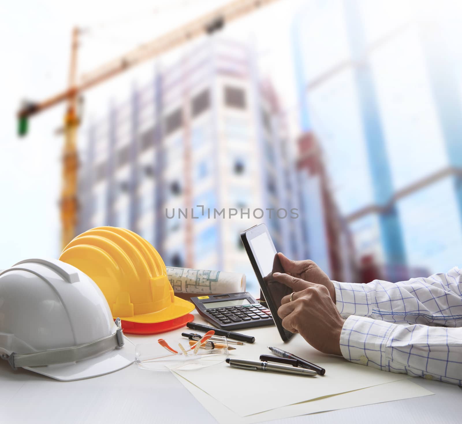 hand of architect working on table with tablet computer and working tool equipment against reflection of office building and crane construction for civil engineering and construction industry business 