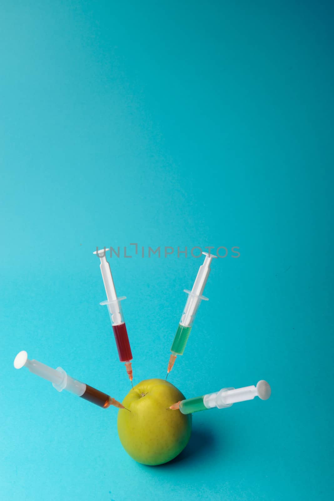 Chemical additives in food or genetically modified fruit concept. Green apple with syringes of chemicals. Isolated on blue background.
