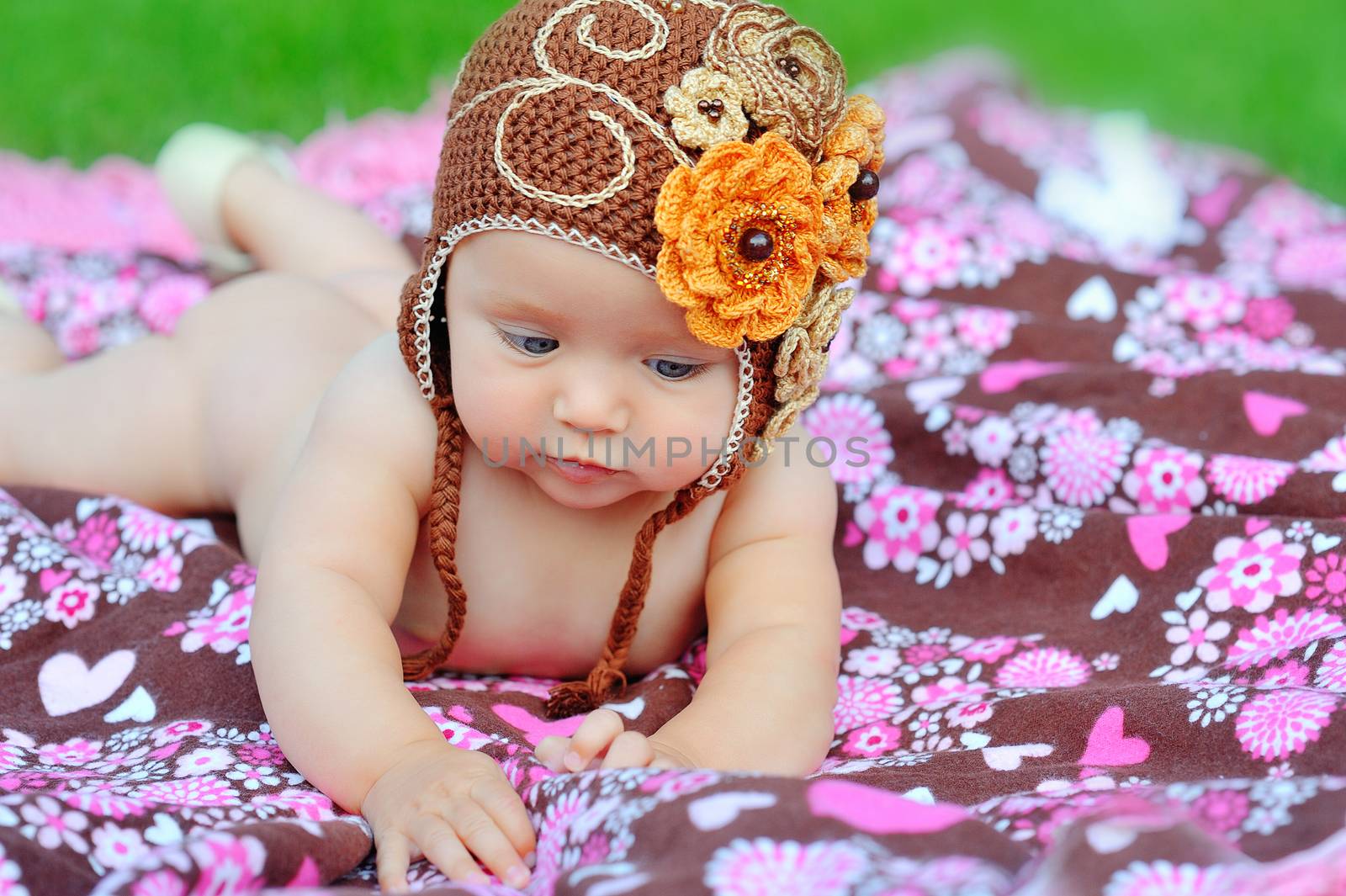 A happy young baby is sitting on green grass outside with bright clouds in the background. 