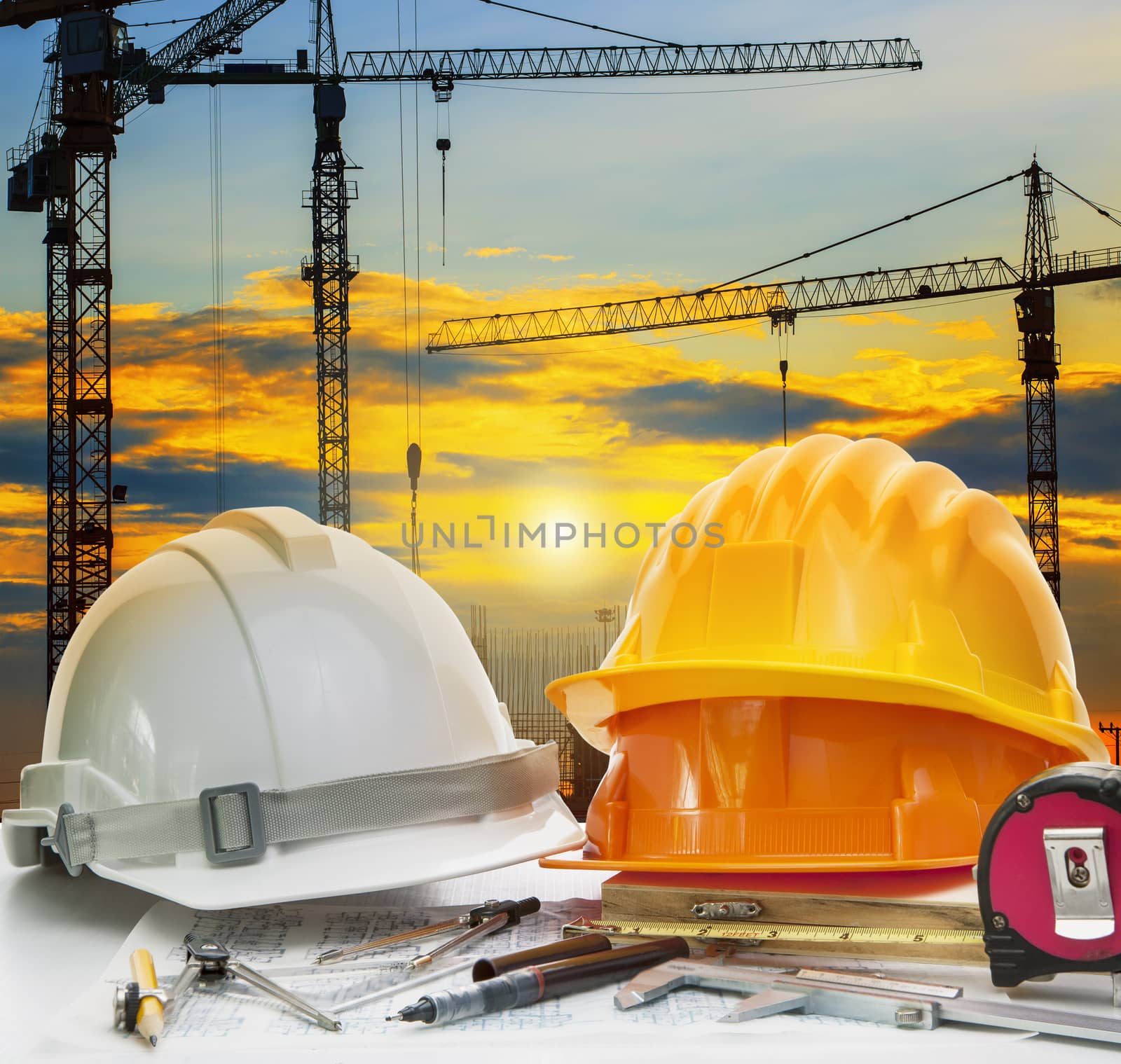 civil engineer working table with safety helmet and writing instrument against beautiful dusky sky and crane construction site