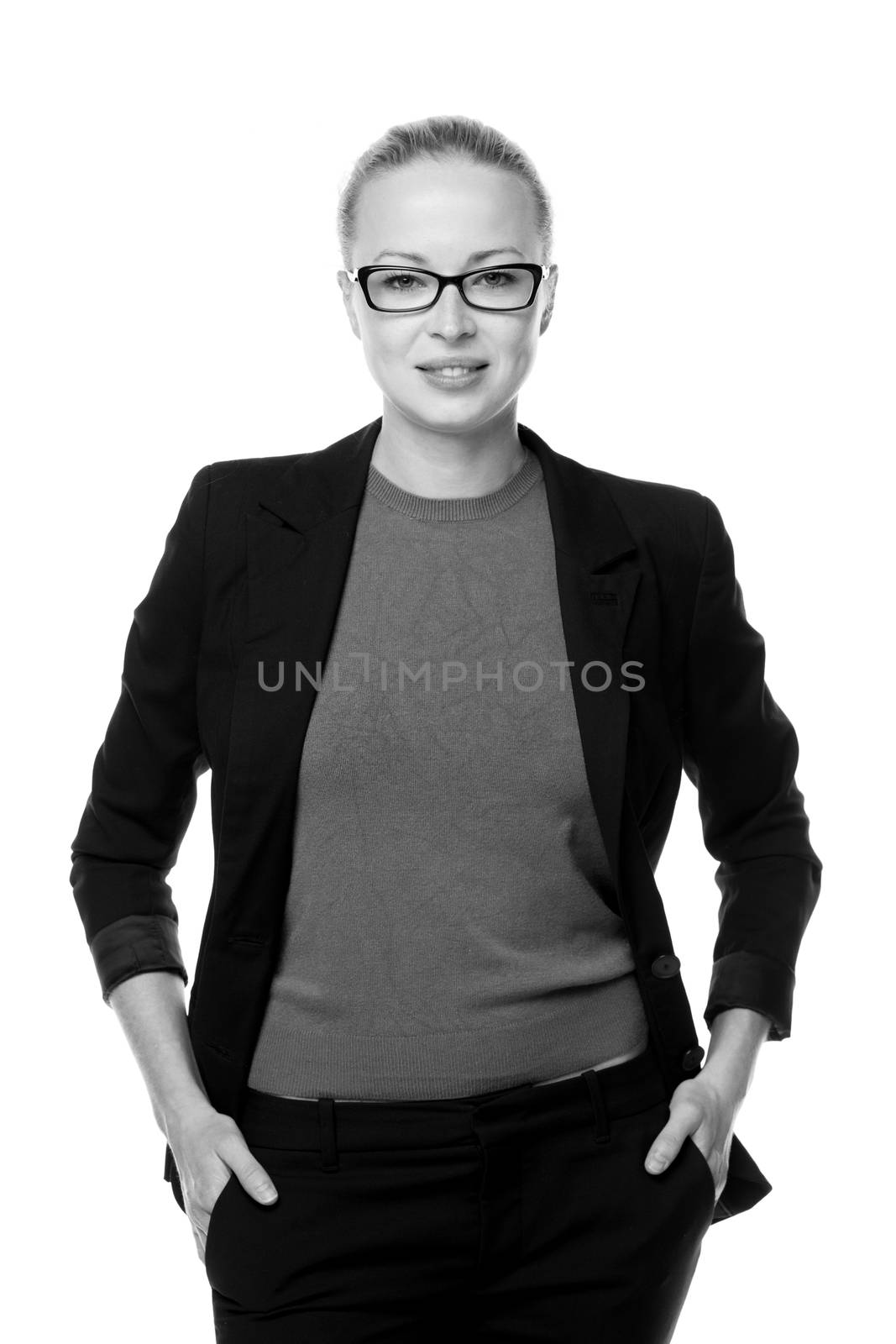 Black and white portrait of beautiful smart young businesswoman in business attire wearin black eyeglasses, standing with arms in pockets against white background.