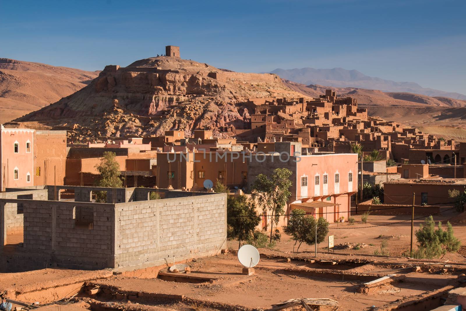 View on a hill with an  old medieval city, built in red color. New building in the foreground, Atlas mountains in the background. Blue morning sky.