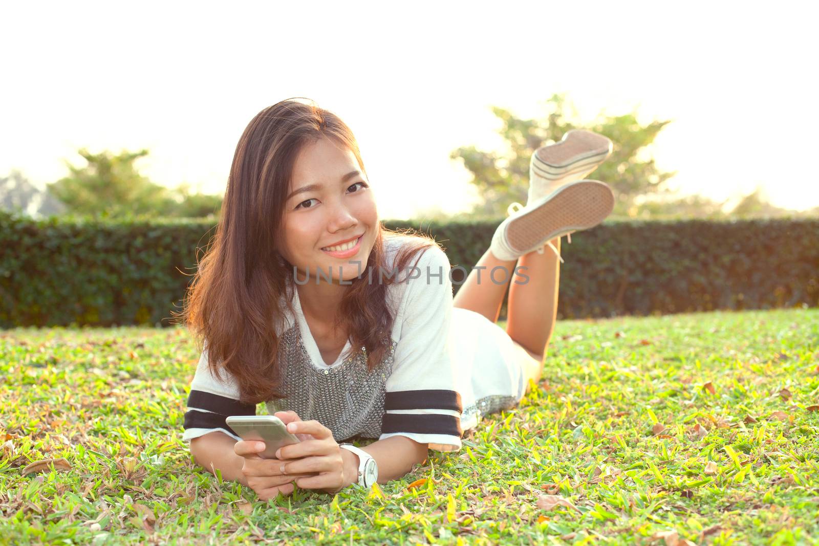 portrait of beautiful young woman lying on green grass field and holding modern smart phone in hand and smiling with happy face emotion