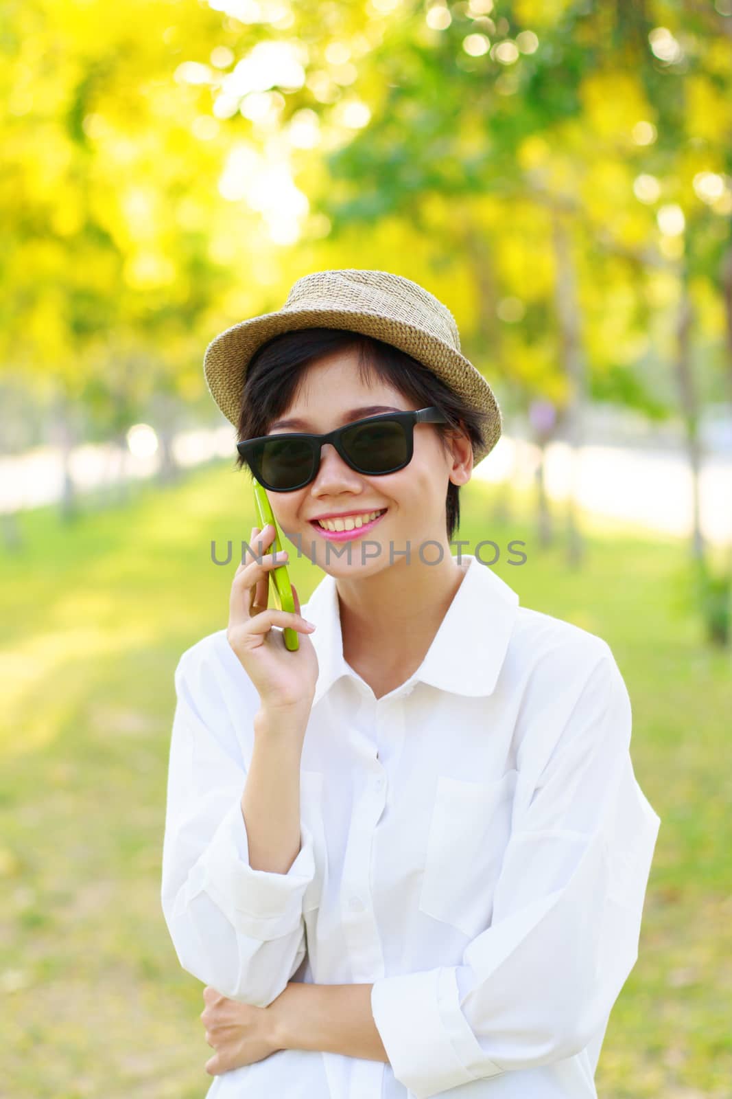 young beautiful woman wearing sun glasses and fashion straw hat talking on mobile phone with happy face toothy smiling against blur of yellow flowers blooming in park