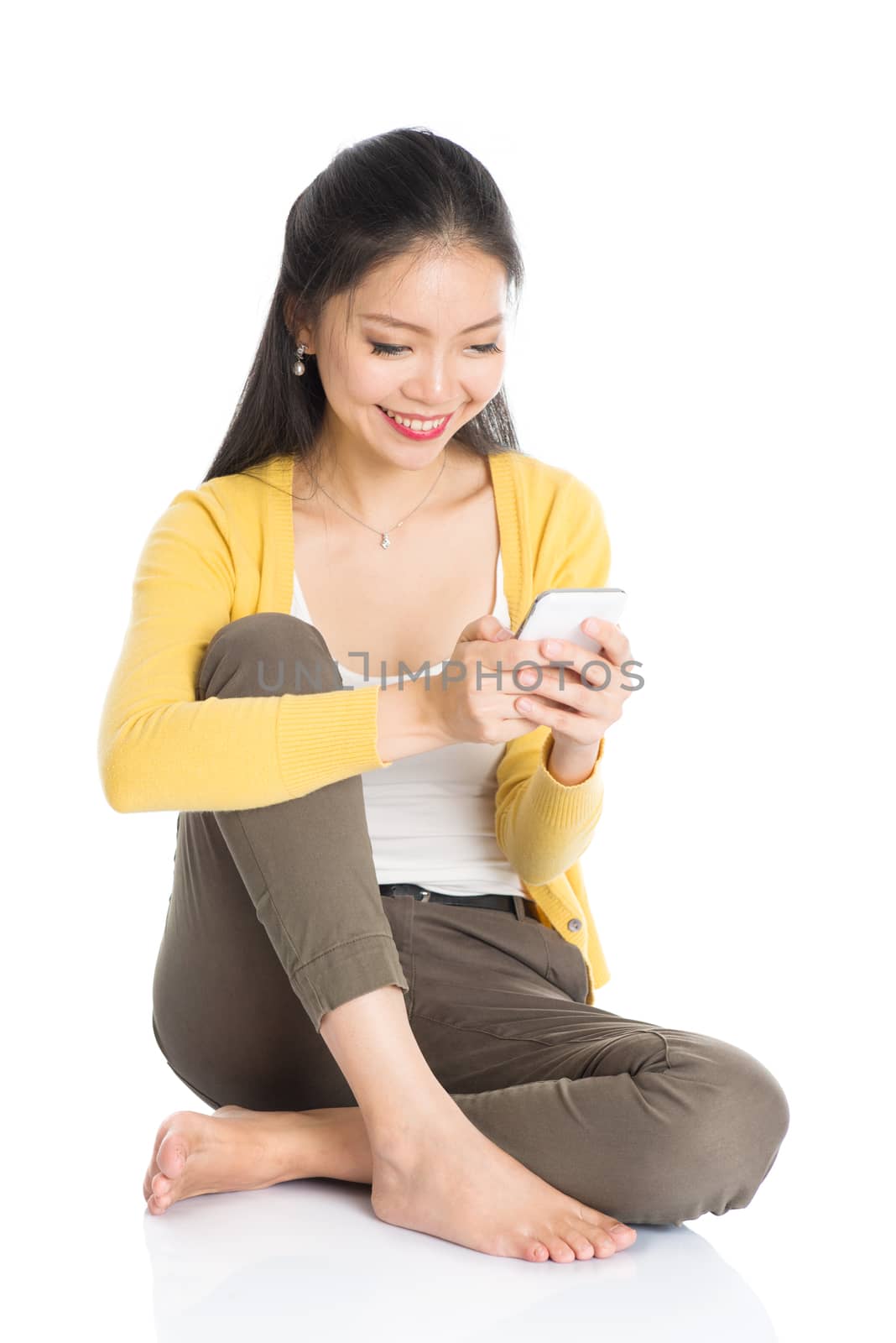 Full length casual Asian girl sitting on floor smiling and using smartphone, isolated on white background.
