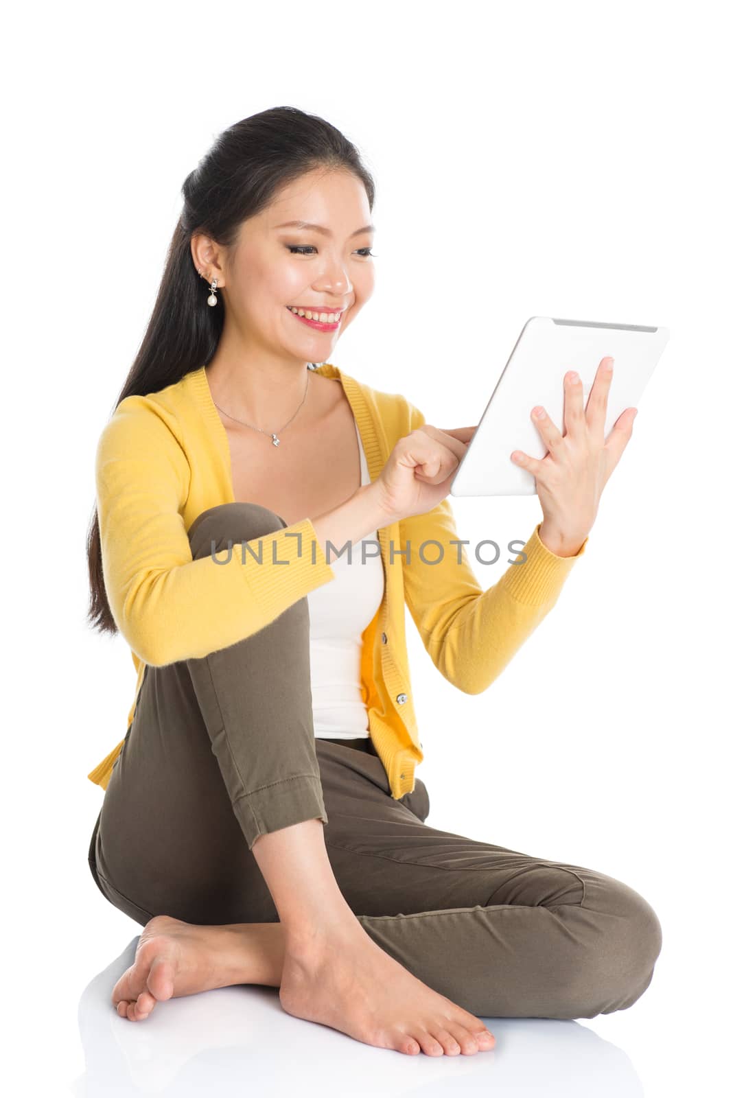 Full length portrait of casual Asian woman sitting on floor smiling and using touch screen tablet pc, isolated on white background.