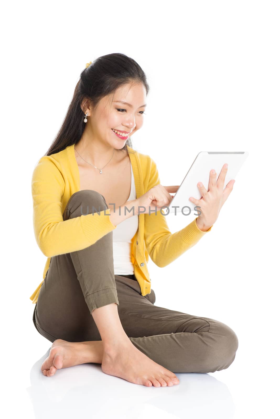 Full length portrait of casual Asian girl sitting on floor smiling and using touch screen tablet pc, isolated on white background.