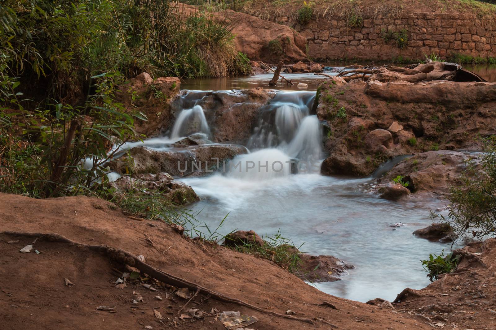 River in the city Cascades d Ouzoud, surrounded by the trees and bushes of an oasis. Dark orange color of the soil. Terrain creating a small waterfall.