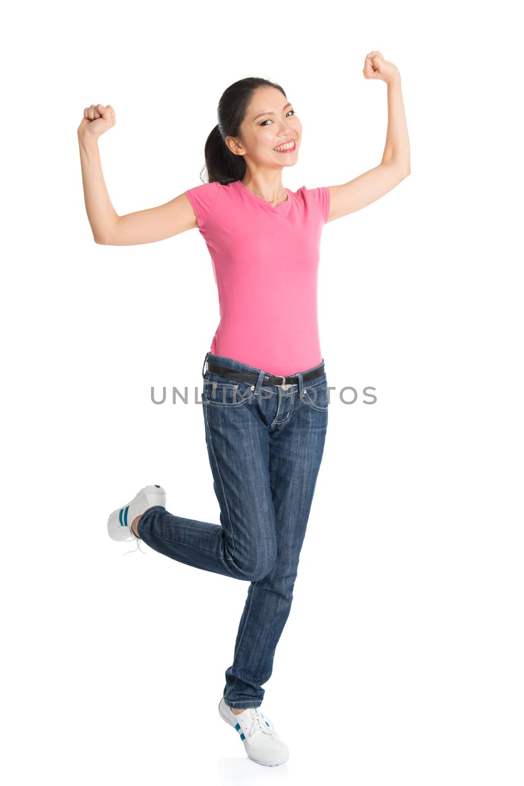 Portrait of excited young Asian woman in pink shirt and jeans arms raised celebrating success, full body standing isolated on white background.