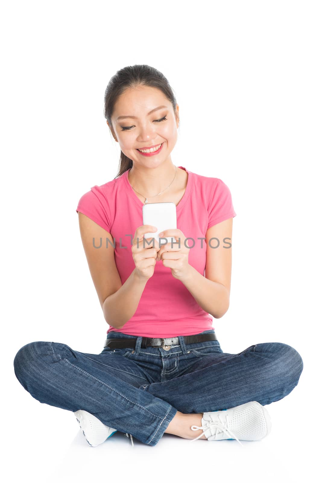 Full body young Asian woman in pink shirt texting with smartphone, seated on floor, full length isolated on white background.