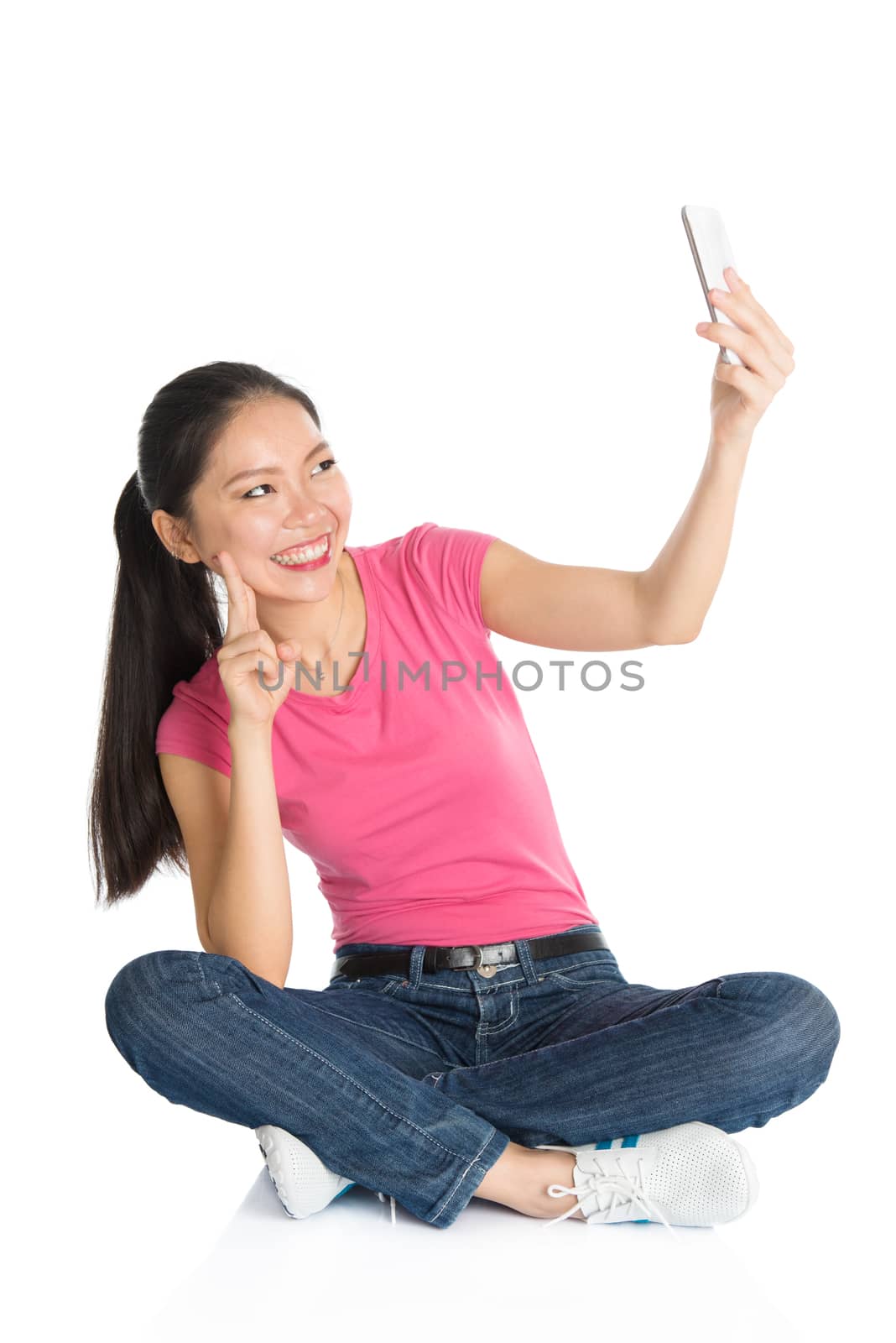 Full body young Asian girl in pink shirt taking self photo or selfie with smartphone, seated on floor, full length isolated on white background.