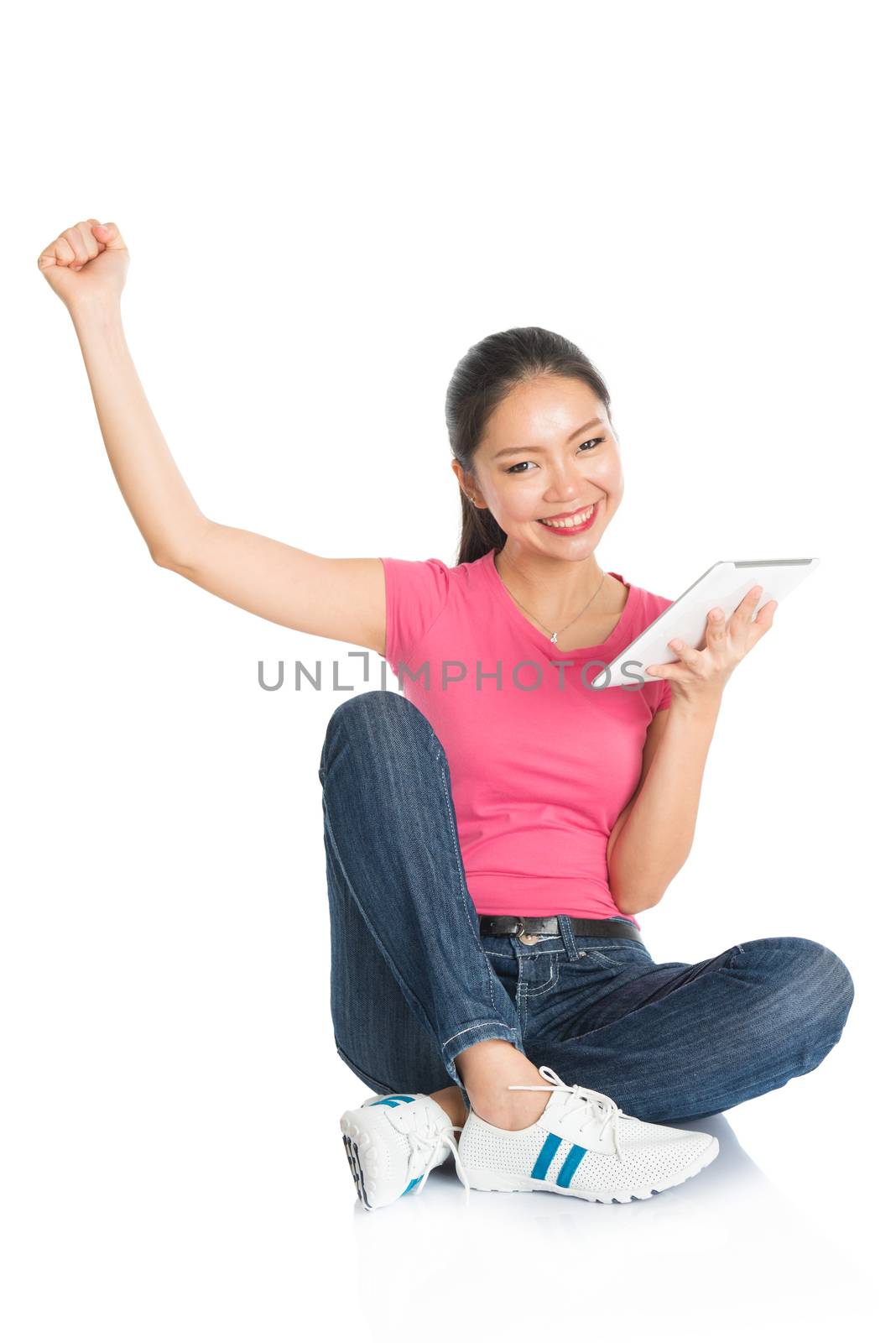 Full body young Asian girl in pink shirt using digital tablet computer and cheering, seated on floor, full length isolated on white background.