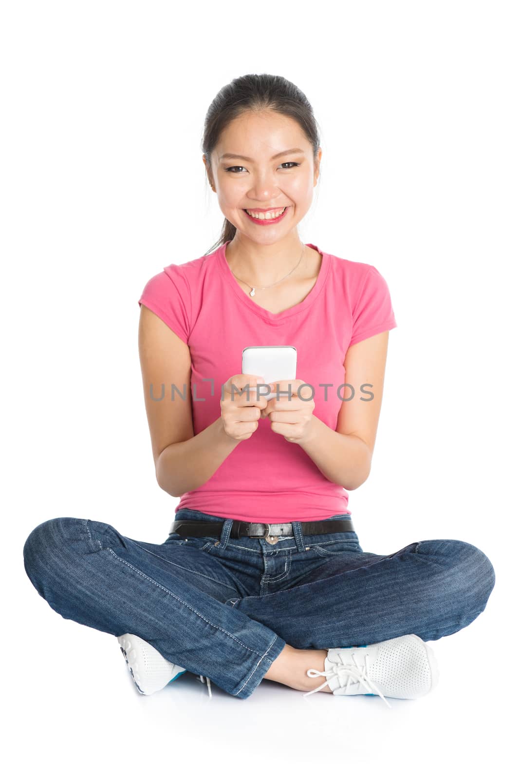 Full body young Asian girl in pink shirt texting with smartphone, seated on floor, full length isolated on white background.