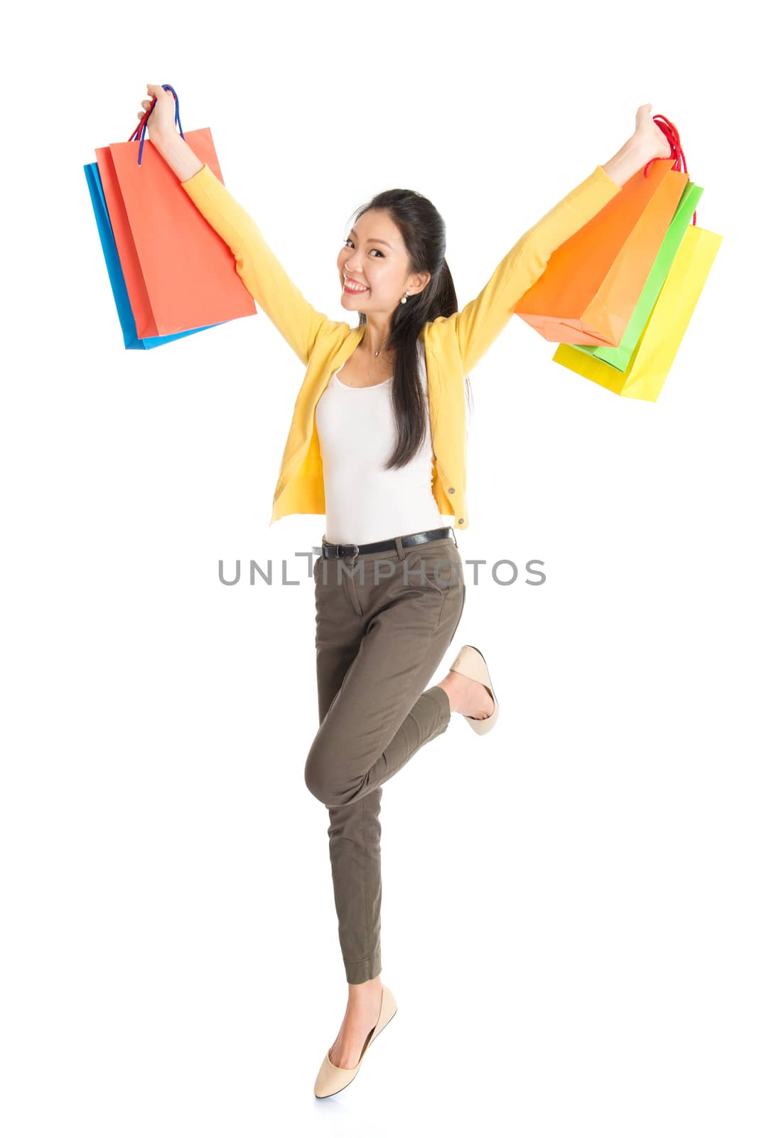 Happy young Asian woman shopper jumping, hands outstretched holding shopping bags and smiling, full length isolated standing on white background.