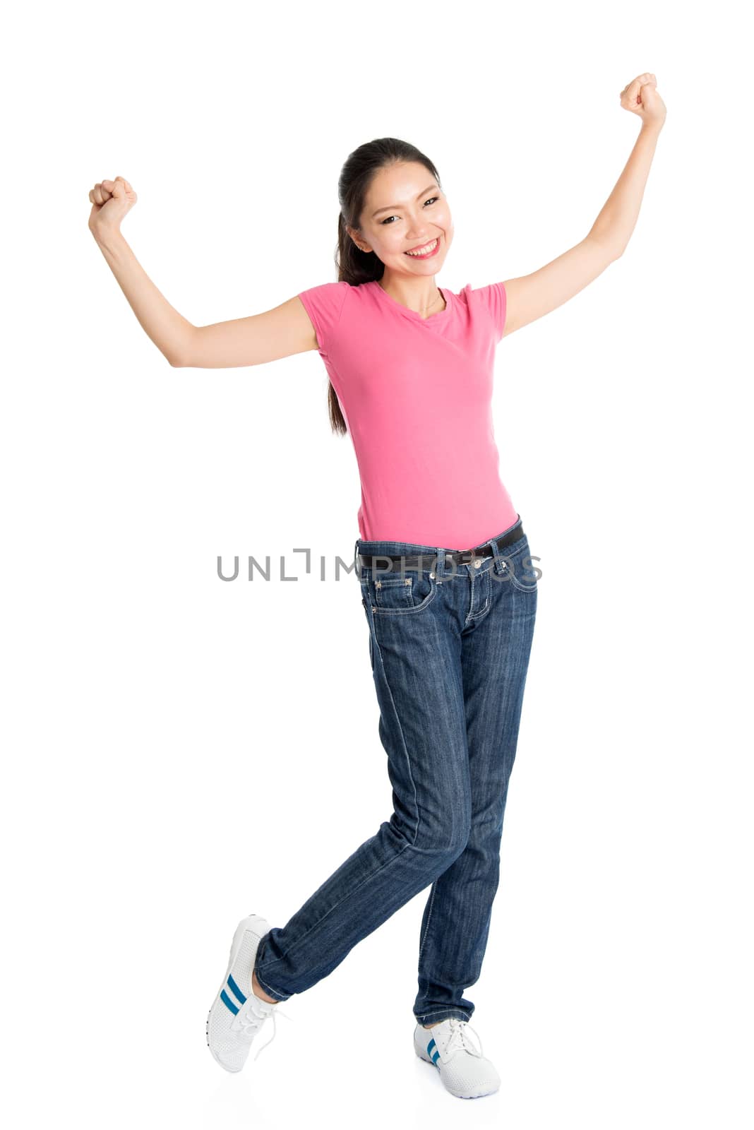 Portrait of excited young Asian girl in pink shirt and jeans arms raised celebrating success, full body standing isolated on white background.