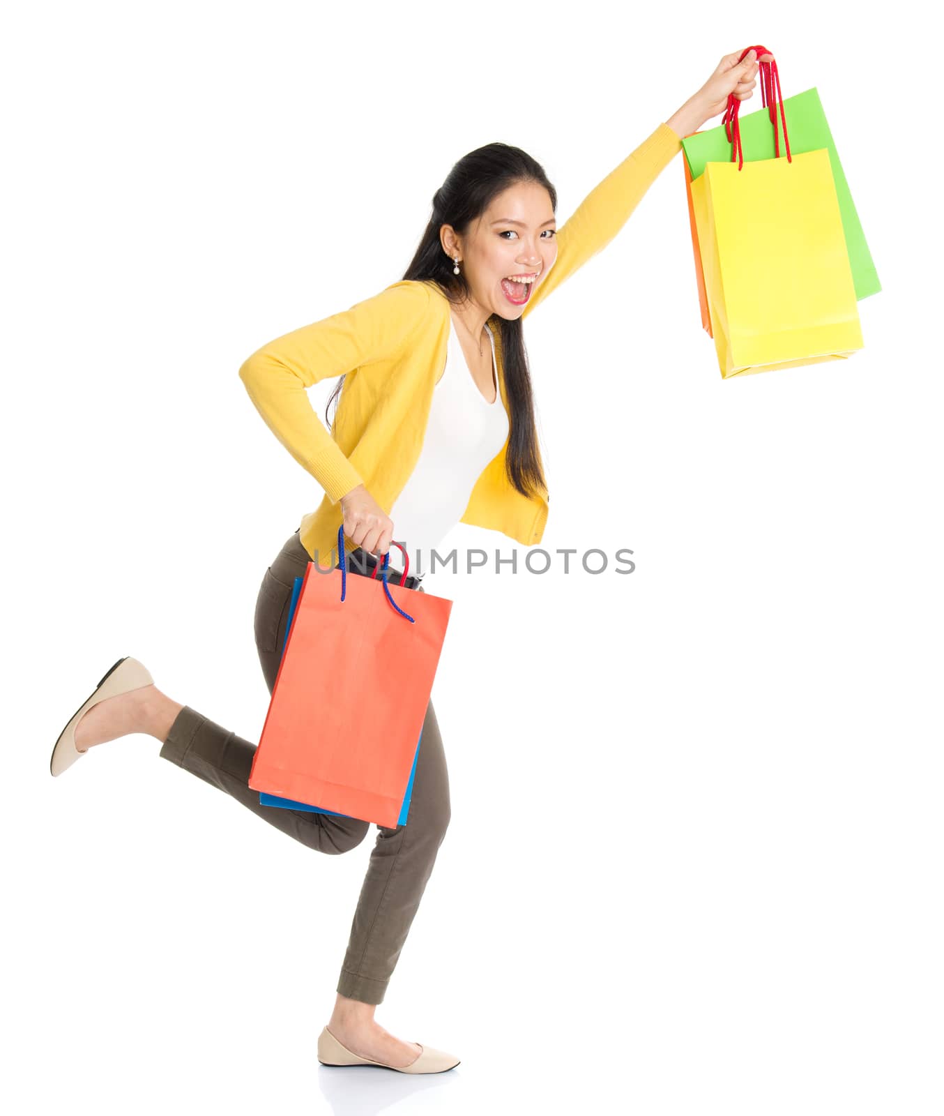 Happy young Asian female shopper running, hands outstretched holding shopping bags and smiling, full length isolated standing on white background.