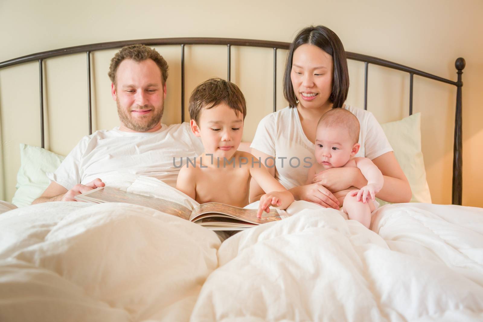 Young Mixed Race Chinese and Caucasian Baby Boys Reading a Book In Bed with Their Father and Mother.