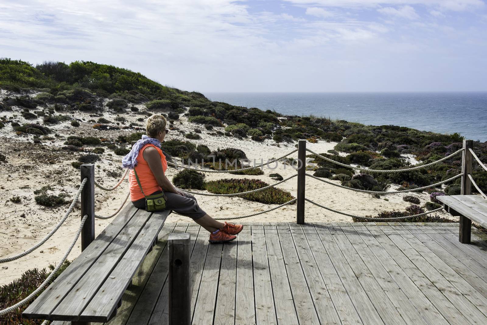 Adult woman with blond hair and orange shirt sitting at the coast of portugal alentejo and looking over the horizon