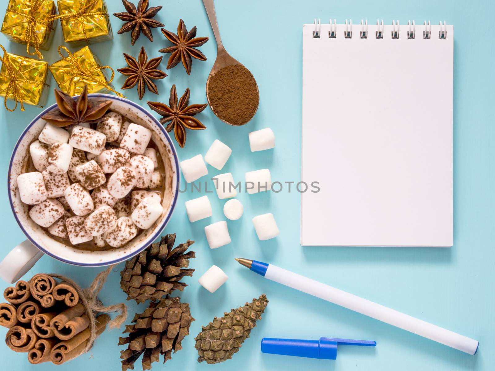 Cup of hot cocoa or carob or chocolate with marshmallow and winter spices star anise and cinnamon sticks, pine cones and carob powder in spoon and gifts and empty notebook on turquoise table. Flat lay