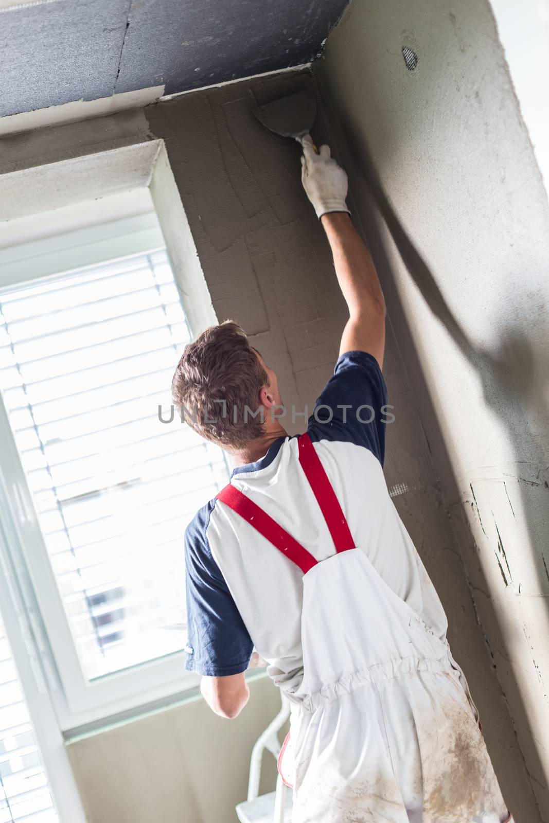 Thirty years old manual worker with wall plastering tools renovating house. Plasterer renovating indoor walls and ceilings with float and plaster.