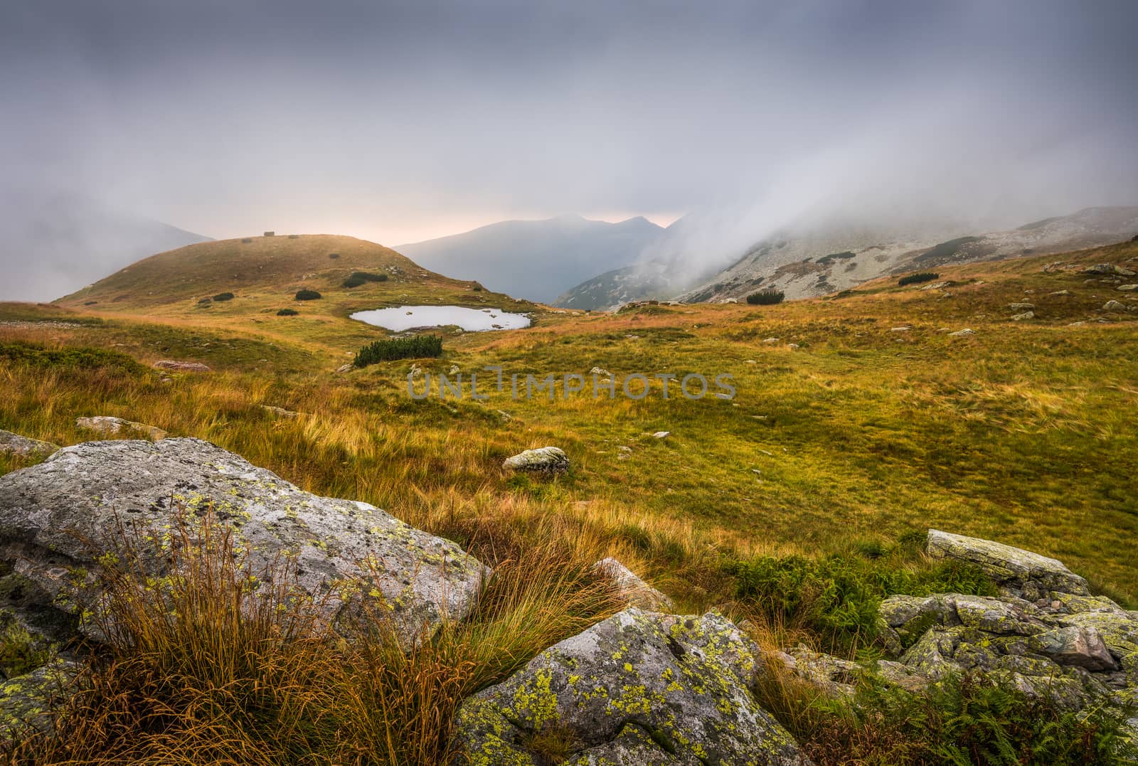 Foggy Mountain Landscape with a Tarn and Rocks in Foreground at Sunset