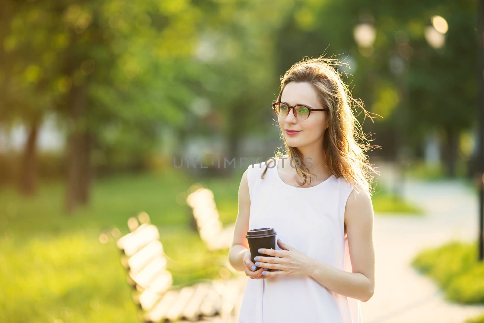 Portrait of lovely urban girl with paper cup in her hands. Happy smiling woman walking in a city park. Fashionable blonde girl wearing spectacles