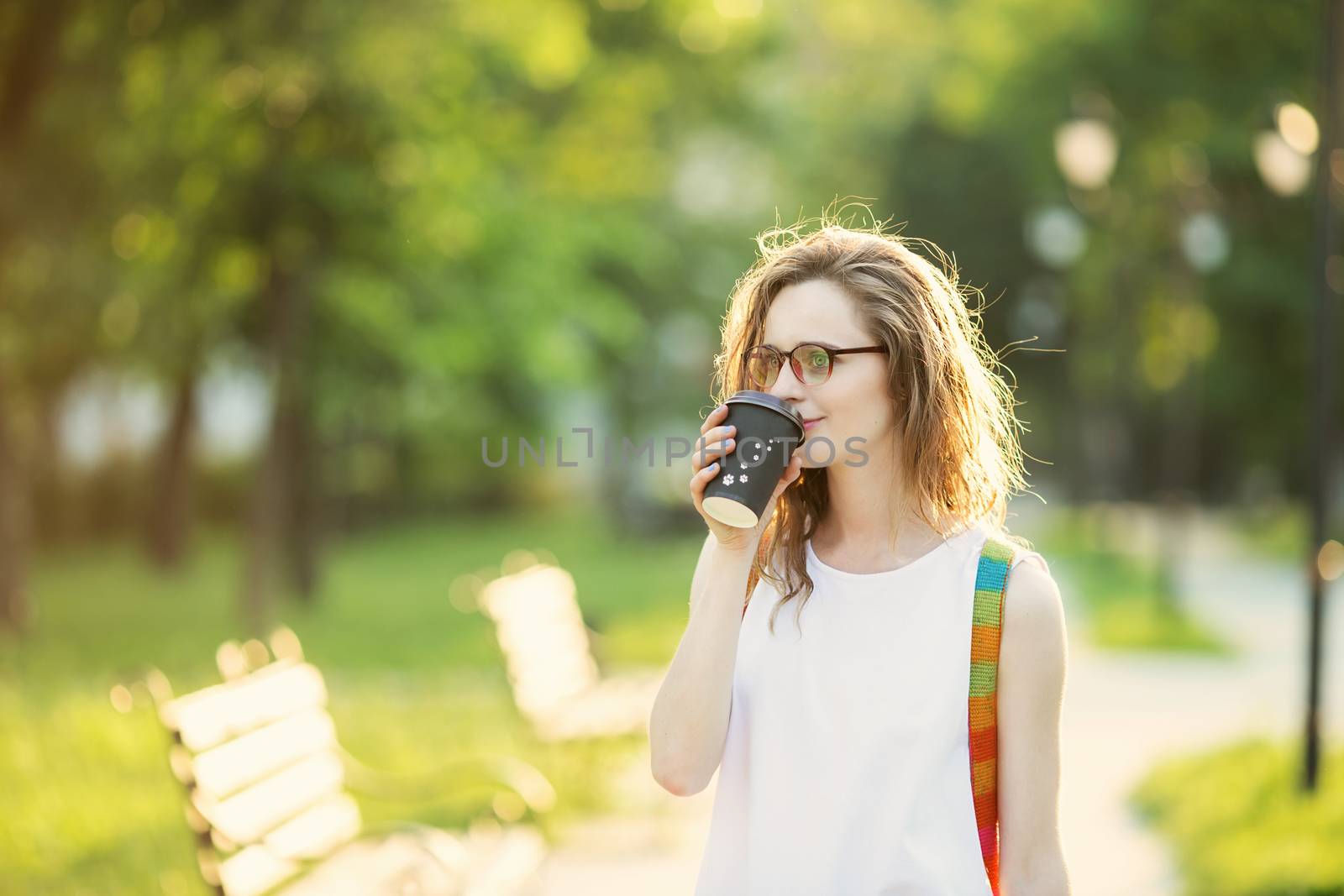 Portrait of lovely urban girl with paper cup in her hands. Happy smiling woman walking in a city park. Fashionable blonde girl wearing spectacles