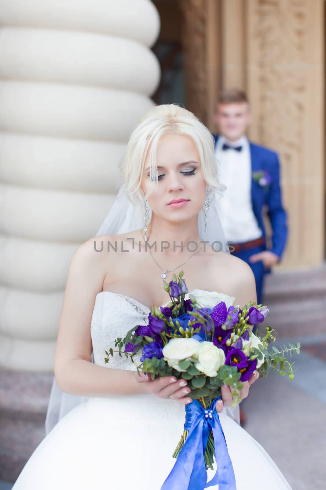 The blonde the bride with a bouquet in hands, on a background the groom in a blue suit. Wedding day.