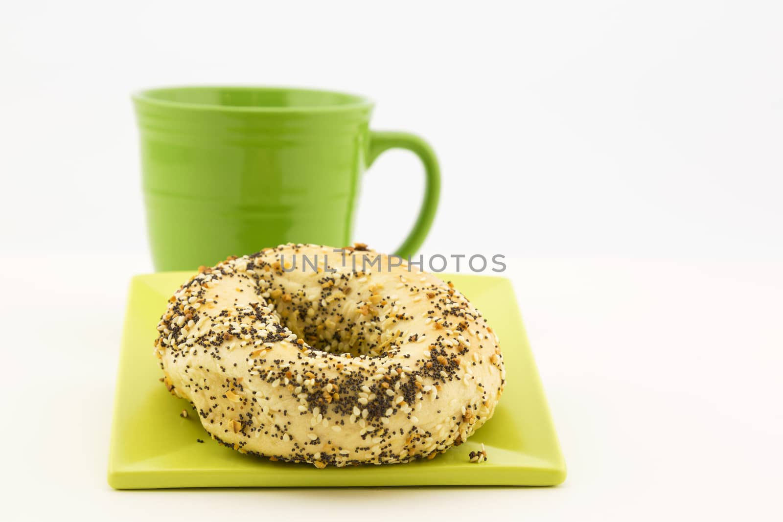 Bagel and coffee presentation with garlic, sesame and poppy seeds as topping and green mug adjacent.  Still life in horizontal image with copy space. Crumbs on plate. 