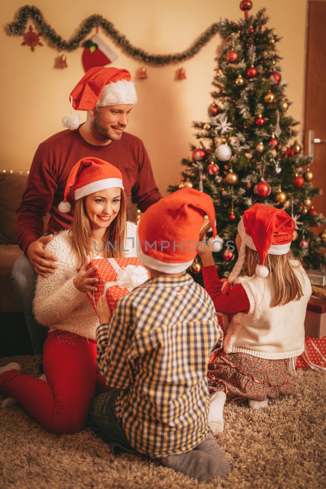 Beautiful happy family wearing santa hat and exchanging Christmas gifts at home. Cute little boy giving a present to his happy young mother. Selective focus.