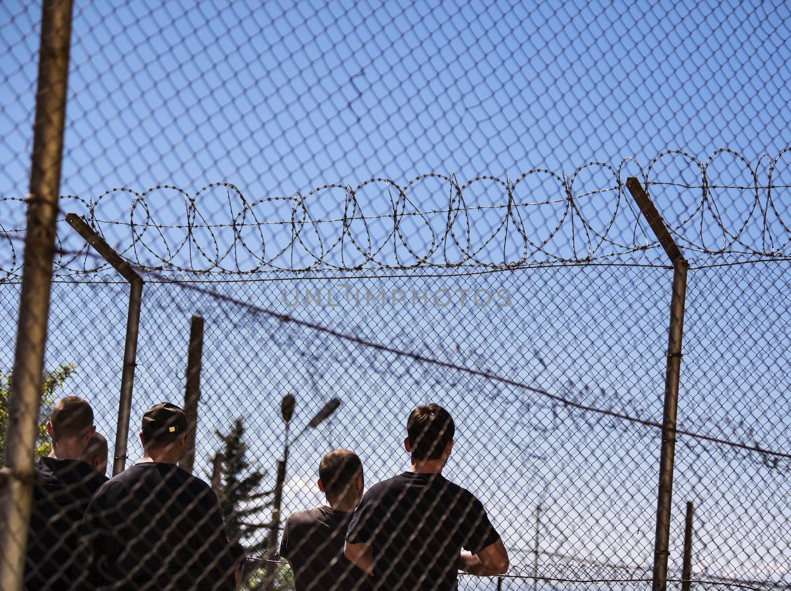 High fence in refugee camp or a jail with razor wire and some people seen from behind.