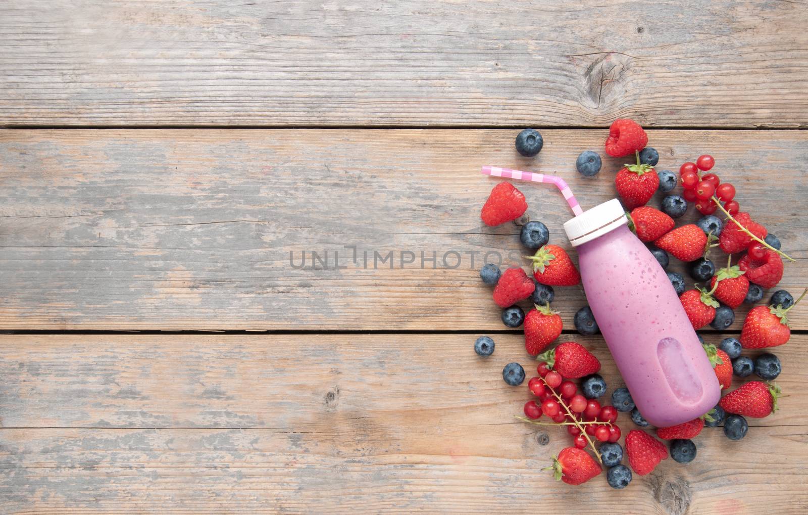 Berry smoothie in a bottle with ingredients on top of a wooden background with space