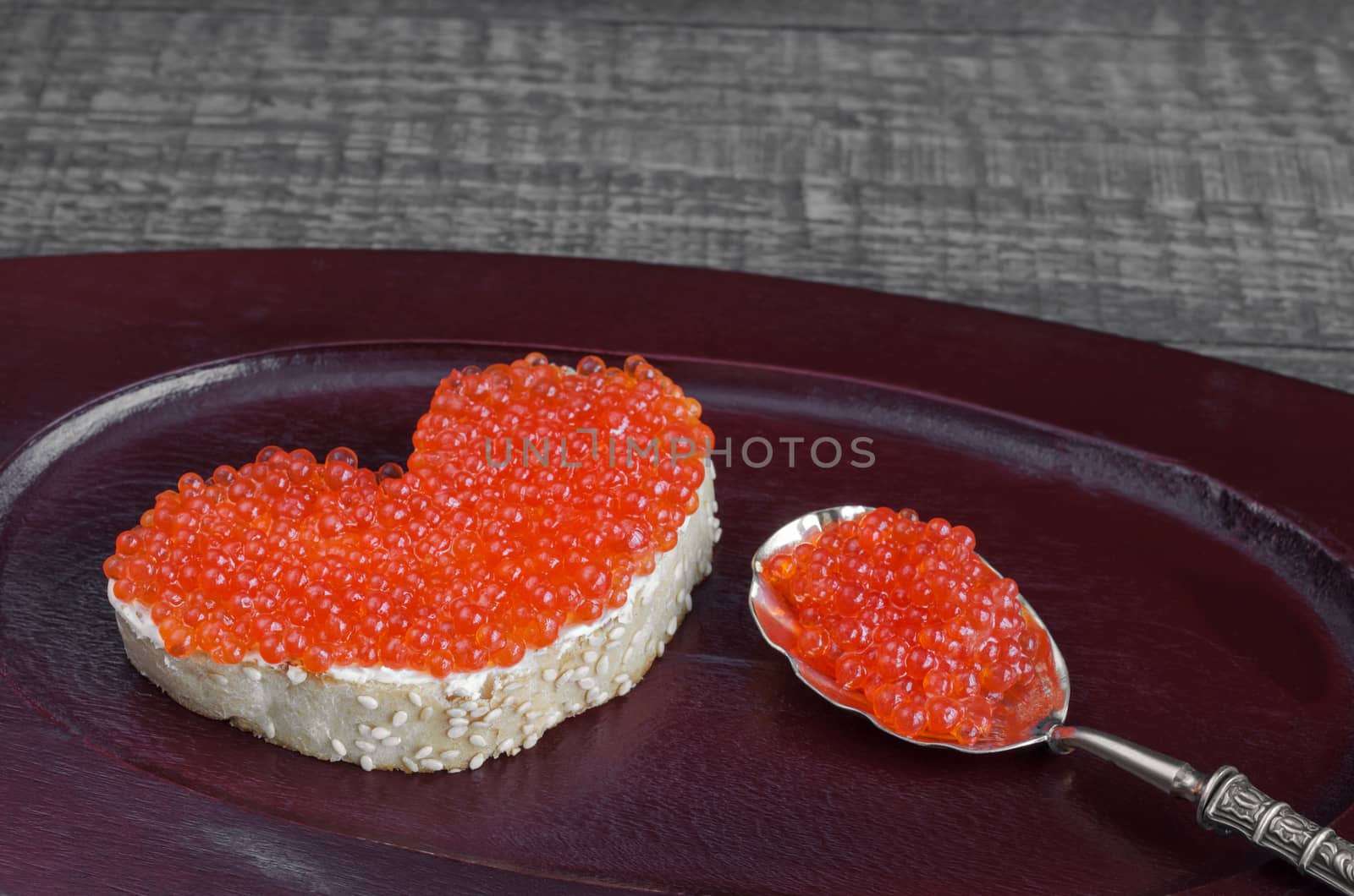 Sandwich with red caviar in a heart shape and spoon on wooden tray and grey background. Analog products.