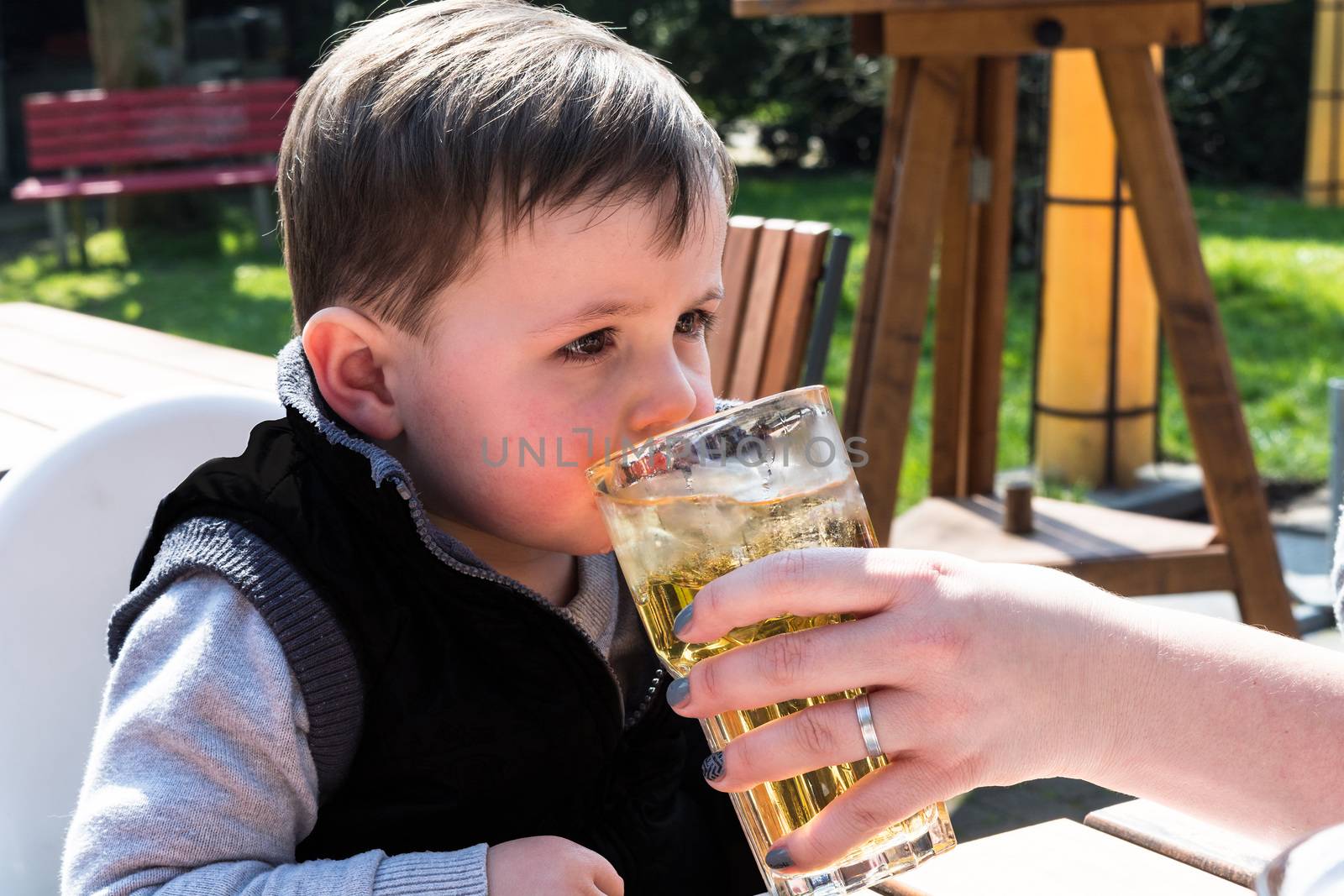 Child drinks juice from a glass in a garden restaurant.
