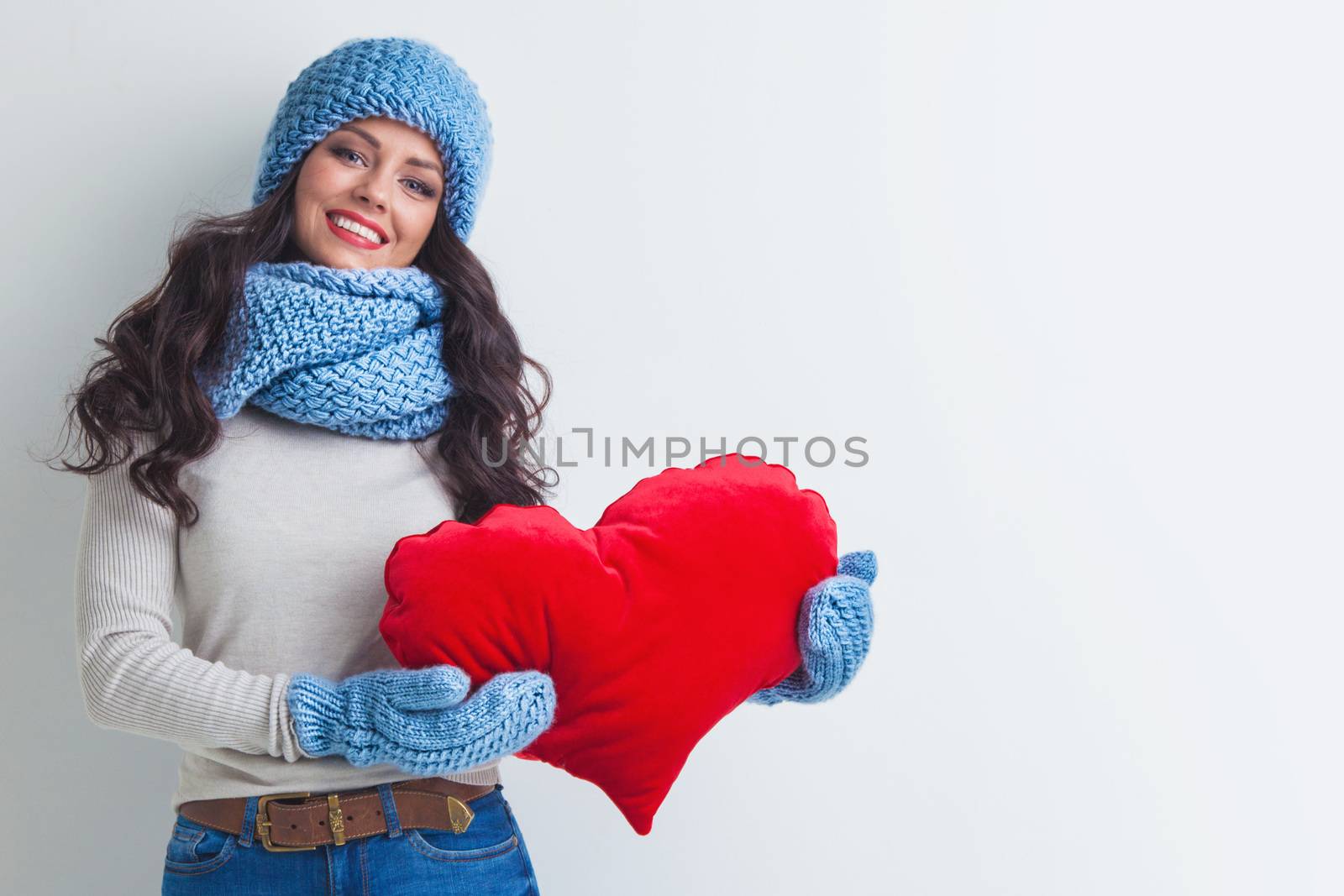 Beautiful smiling brunette woman in winter hat and scarf holding a red heart pillow