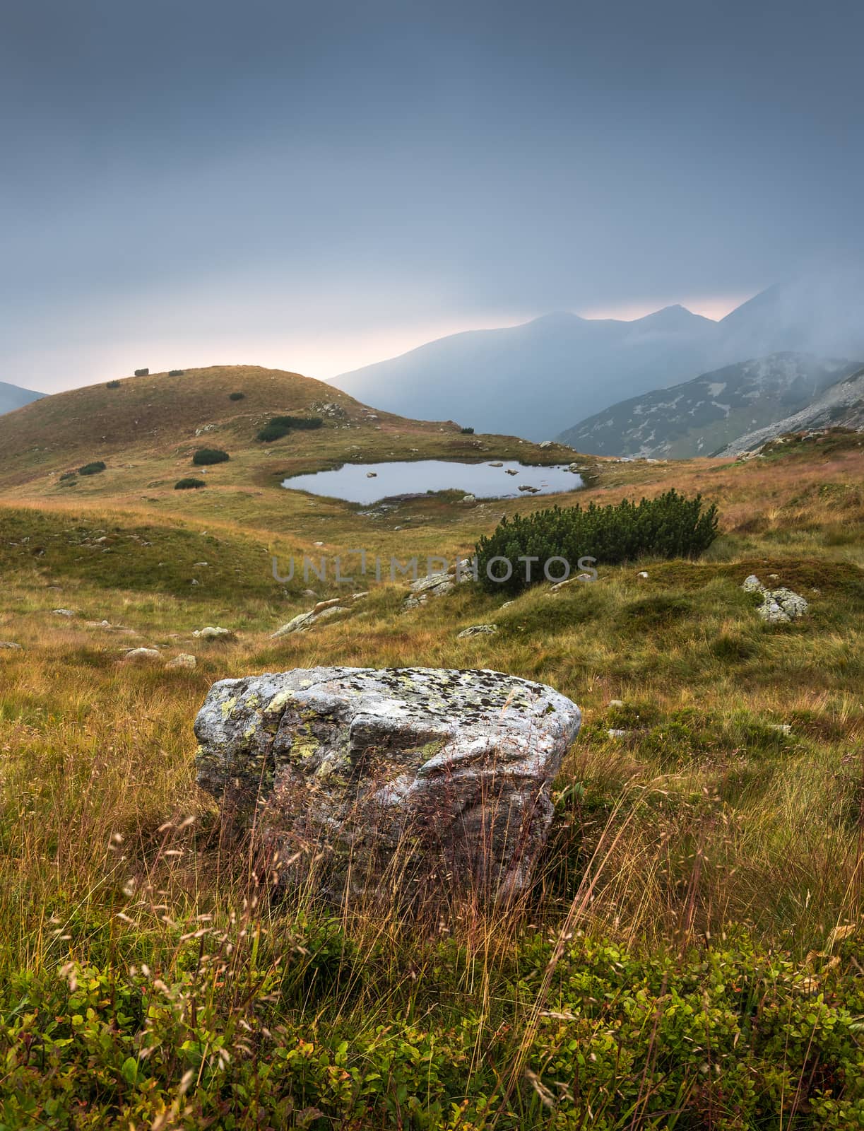 Foggy Mountain Landscape with a Tarn and a Rock in Foreground at Sunset