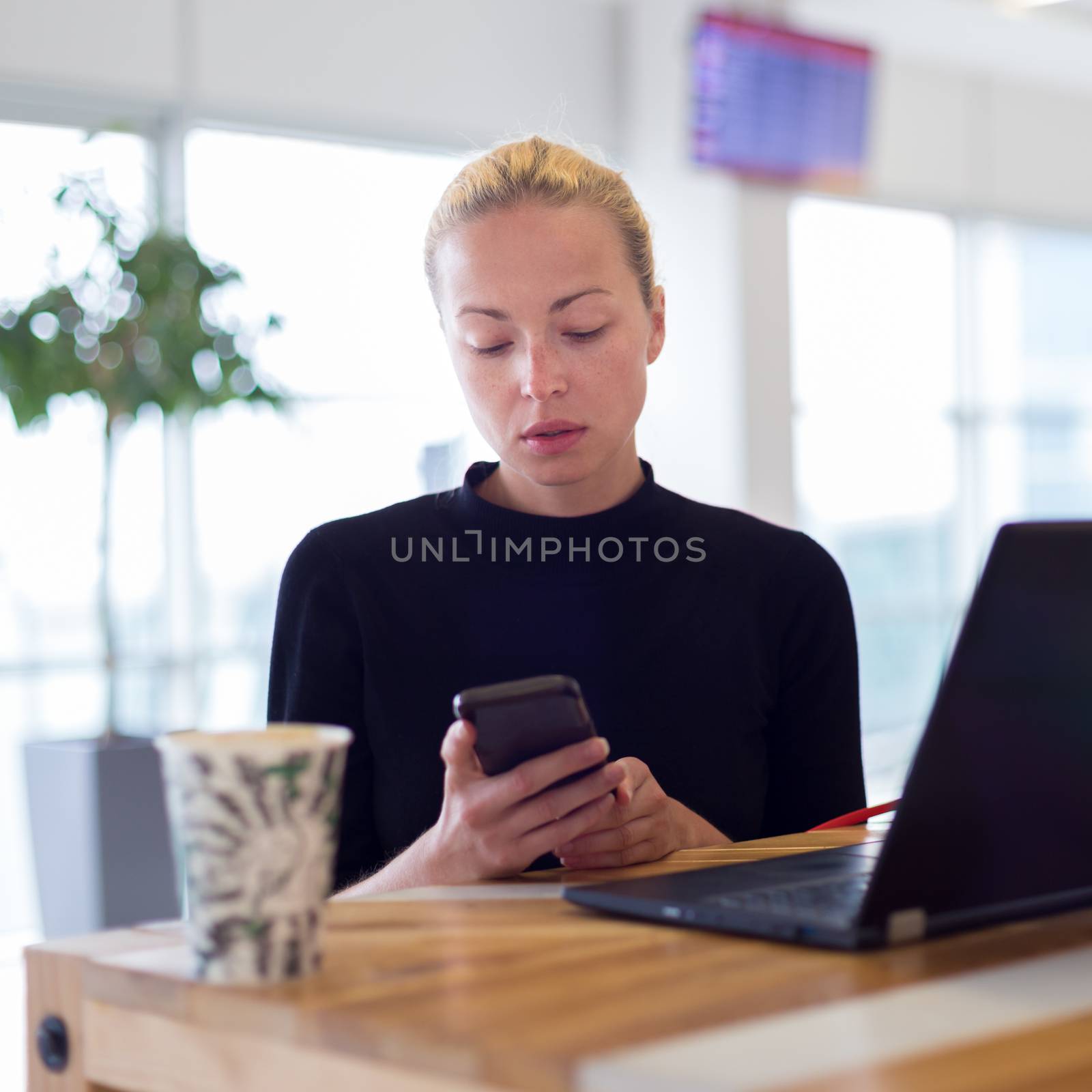 Female freelancer remotely working on the go using multiple devices on a desk at airport coffee shop while traveling. Contemporary entrepreneurial lifestyle.