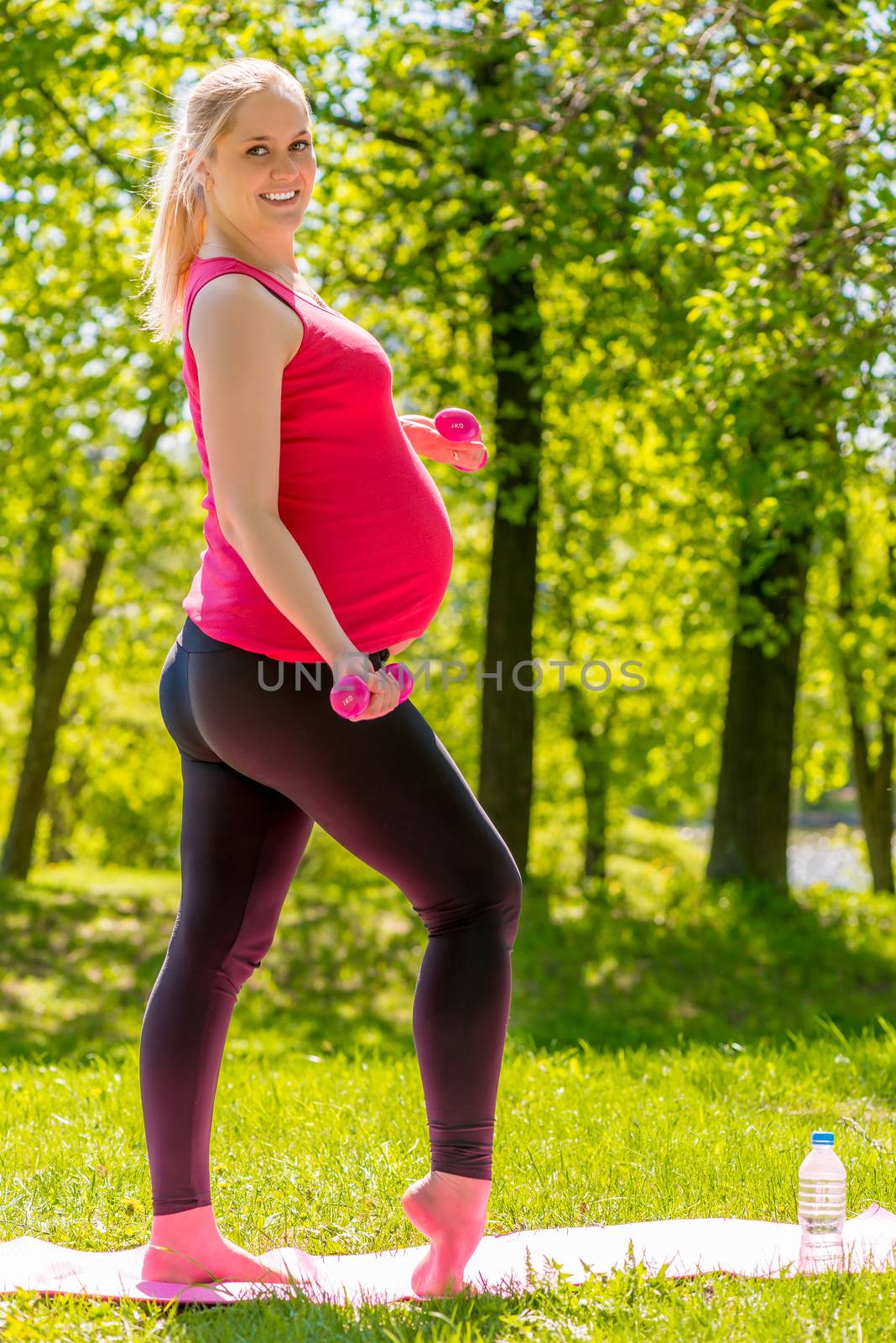 Smiling pregnant woman with dumbbells engaged in sports in the summer park