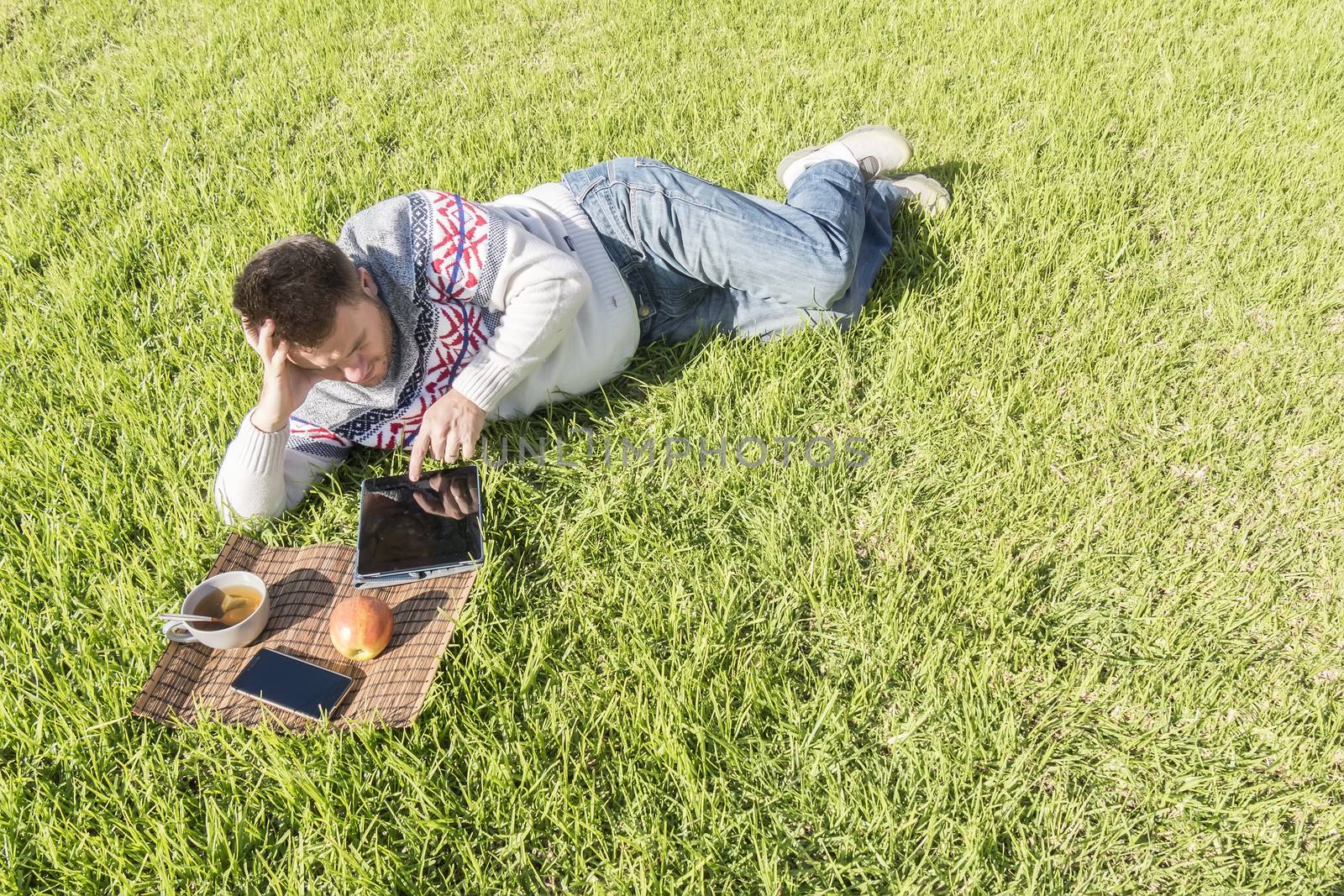 Man having breakfast lying on the grass in the garden with technology