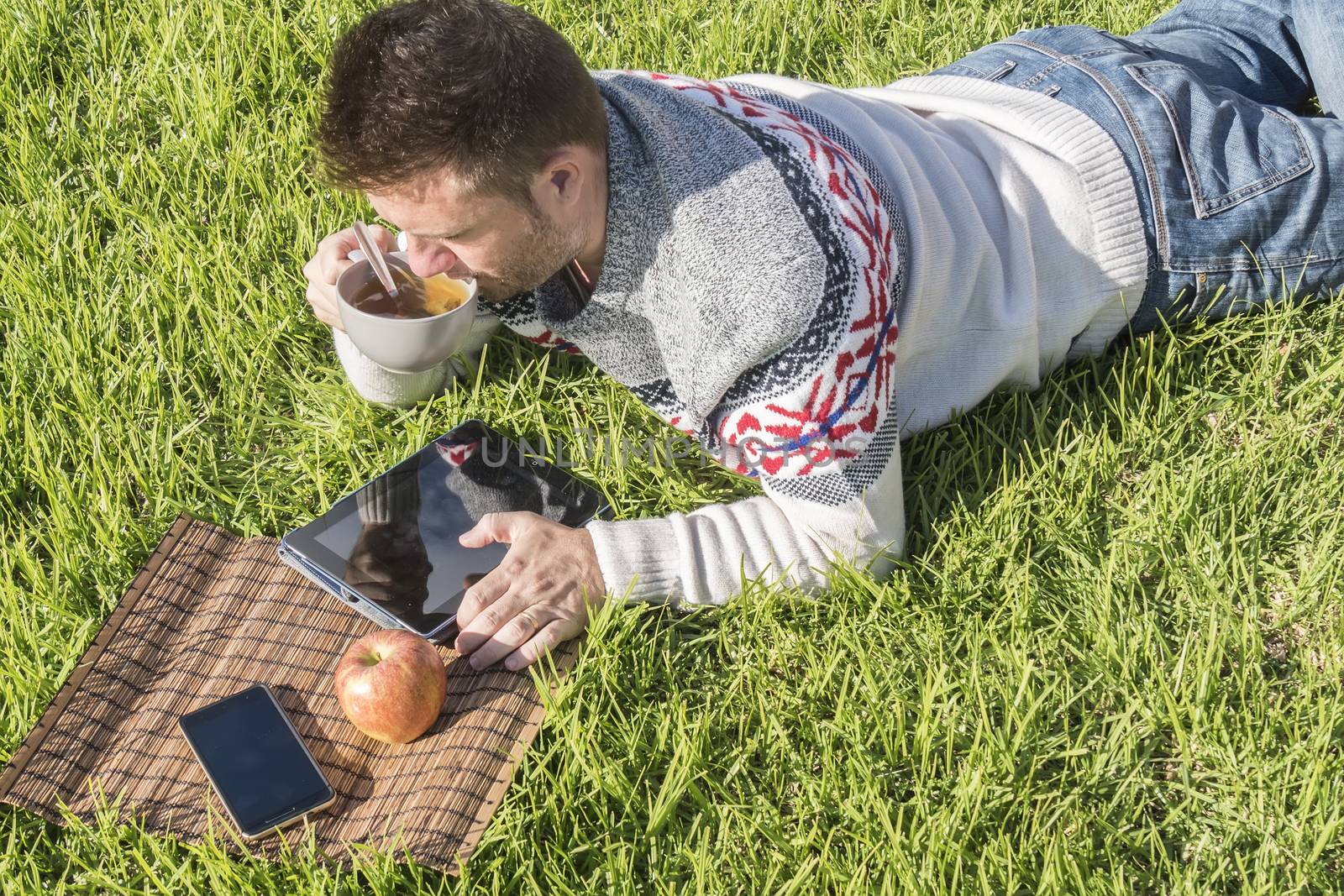 Man having breakfast lying on the grass in the garden with technology