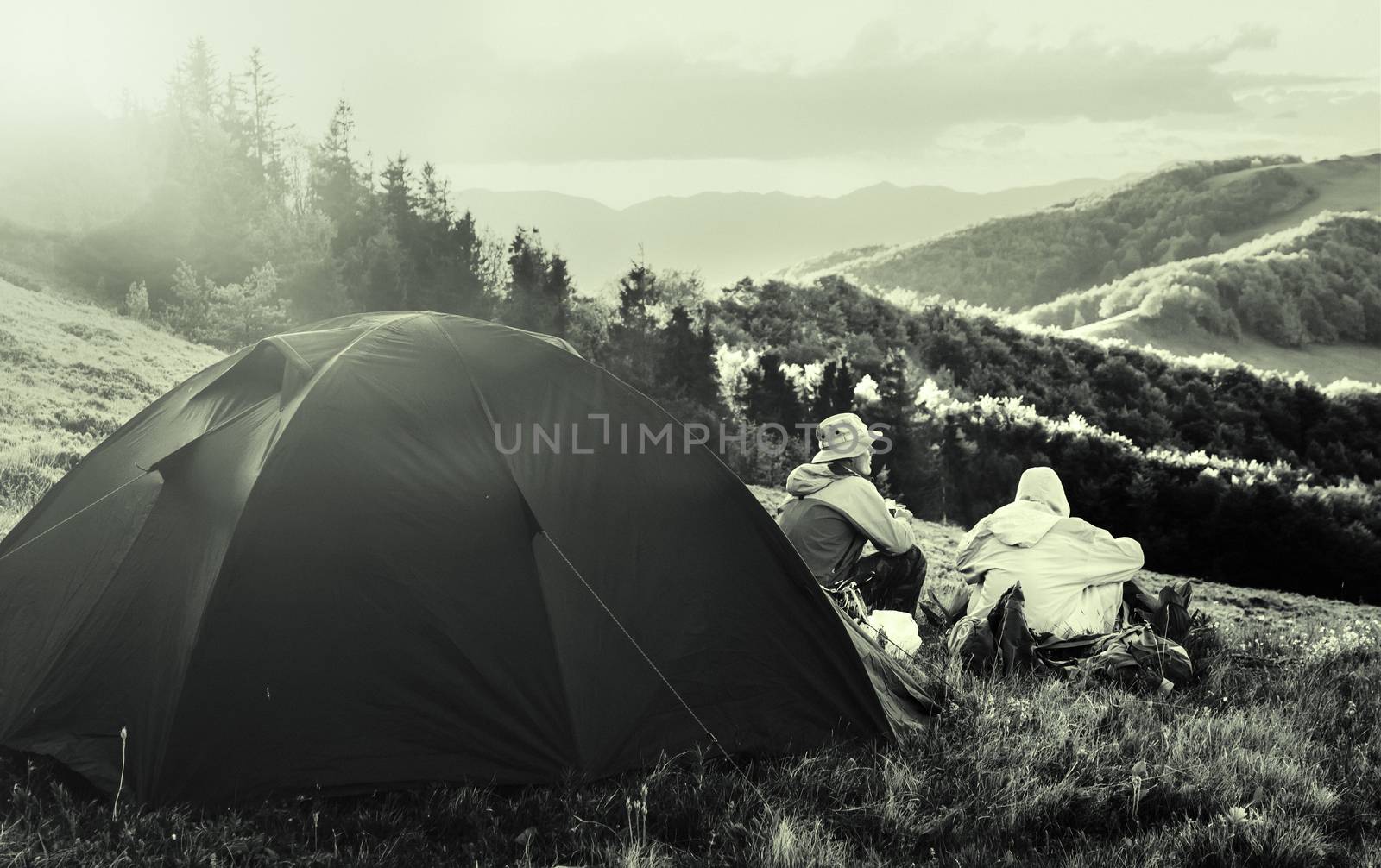 Black and white travel photo. Two tourists sitting near a tent and looking at the scenery. Sun is shining on a tent. Vintage effect. Added noise
